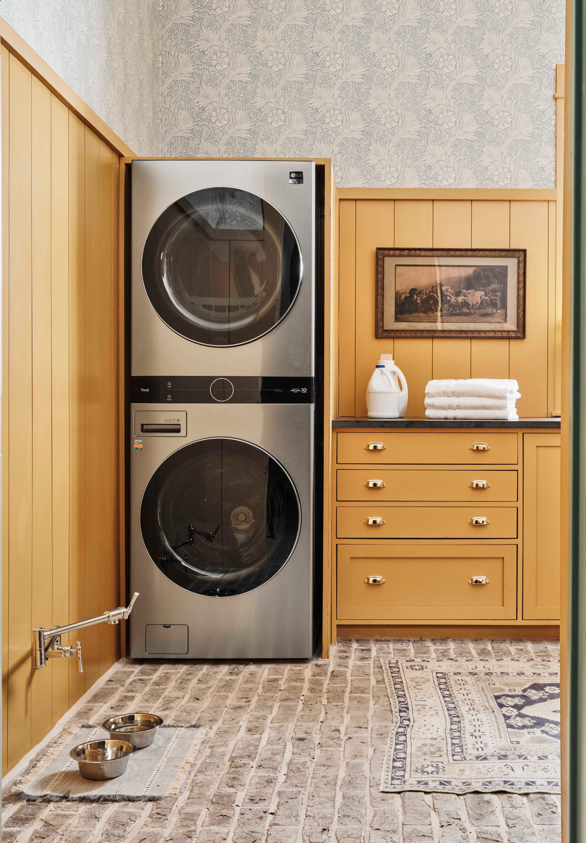 A laundry room featuring a stacked stainless steel washer and dryer, mustard yellow wood paneling, and a rustic brick floor.