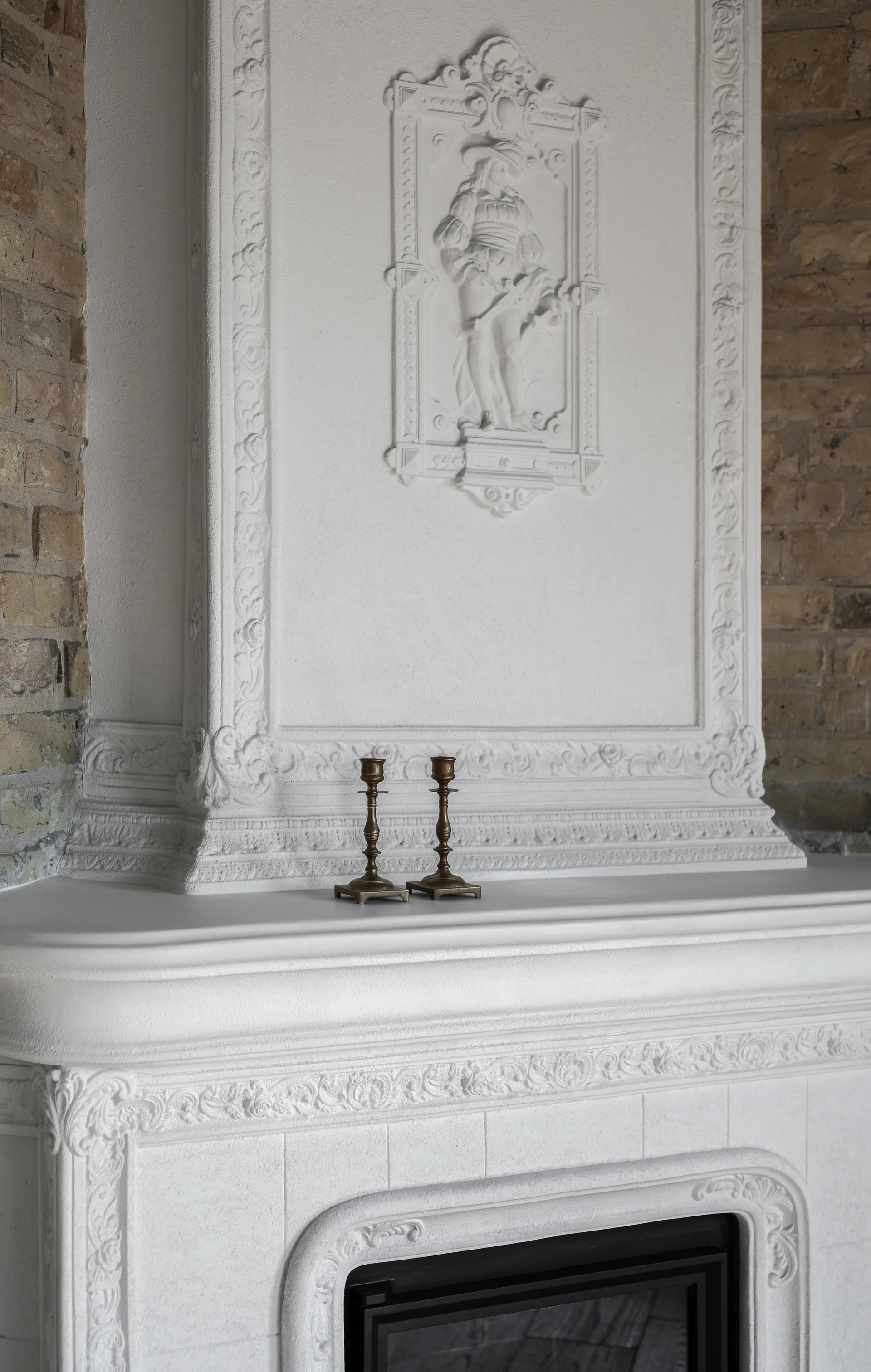 A close-up of an ornate white plaster fireplace with neoclassical relief work, flanked by exposed brick and topped with two brass candlesticks.