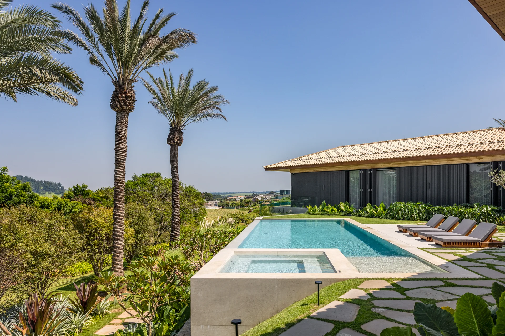 A rectangular backyard swimming pool and integrated spa sit next to a modern home with dark siding, surrounded by flagstone pavers and palm trees.