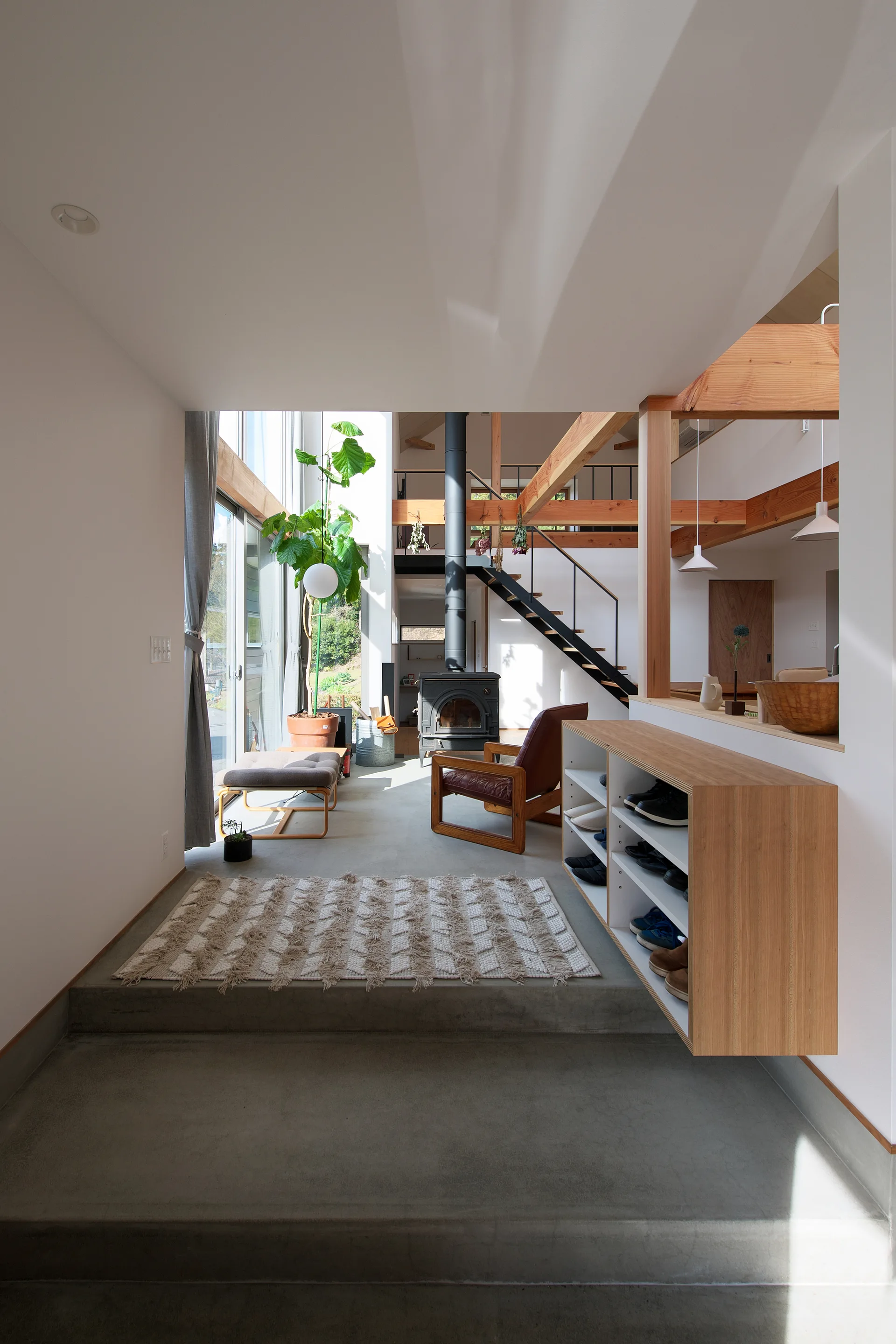 A split-level concrete entryway features a floating wooden shoe cabinet, a patterned rug, and a view of an open-plan living room with wooden beams.