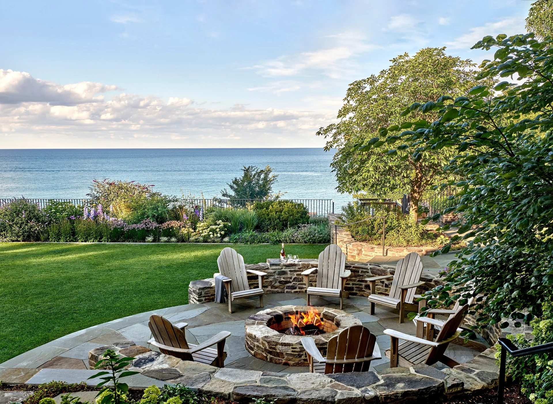 A circular flagstone patio features a stone fire pit surrounded by wooden Adirondack chairs overlooking a green lawn and the ocean.