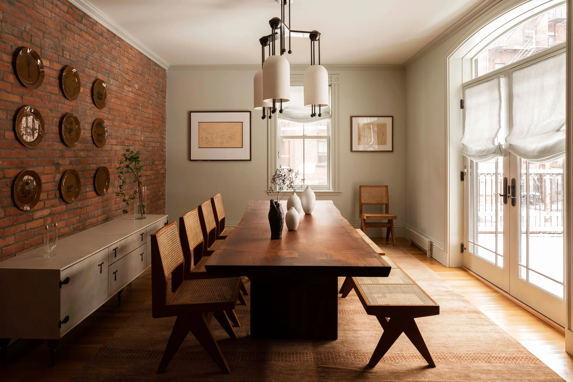 A dining room featuring a long wooden slab table with cane chairs and a bench on a patterned rug, set against an exposed brick wall.