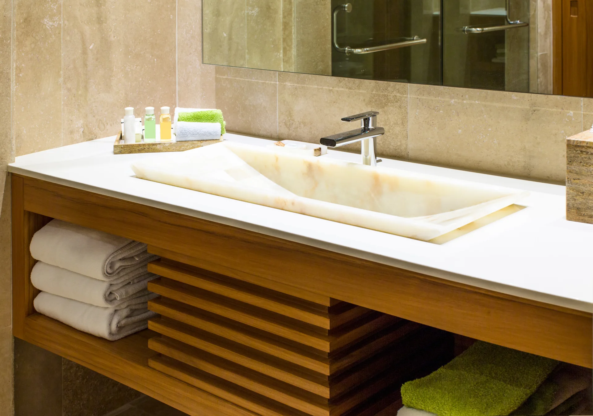 A modern bathroom vanity features a shallow stone vessel sink on a white countertop with a warm wood slatted base and folded towels.
