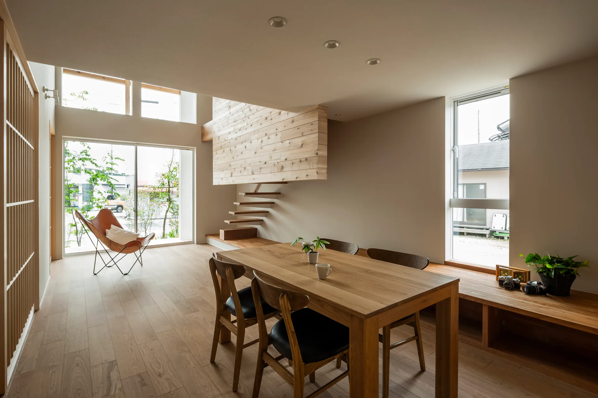 A minimalist dining room features a wooden dining table with chairs, a floating timber staircase, and a tan leather butterfly chair by a large window.