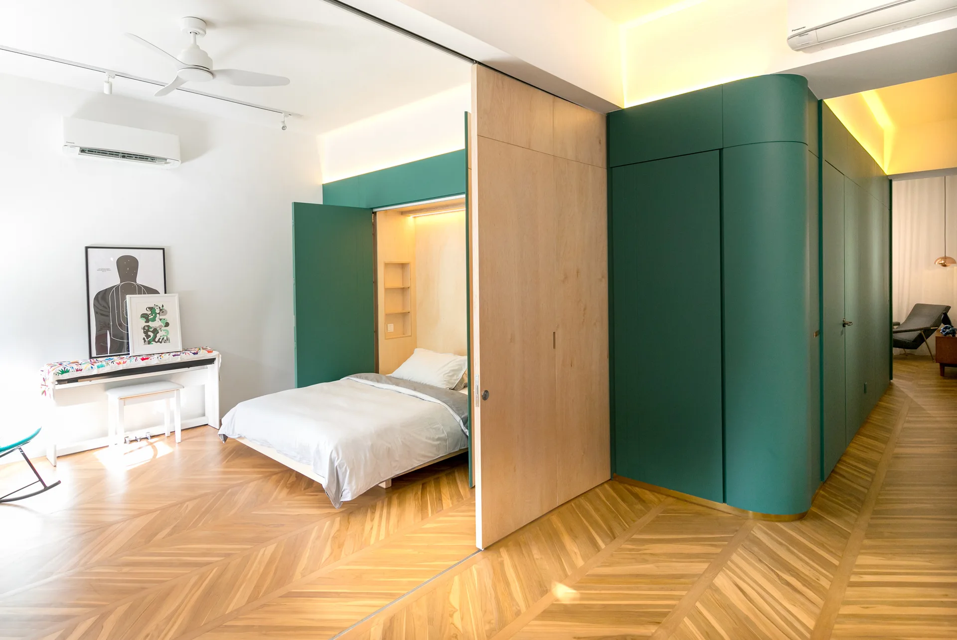 A modern bedroom features a pull-out bed integrated into green built-in cabinetry, herringbone wood floors, and a light wood sliding partition.