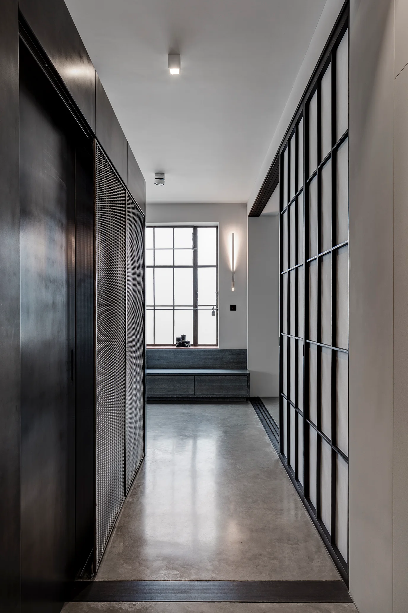 A minimalist hallway featuring polished concrete floors, dark metal-mesh cabinets, and a grid-patterned glass sliding door.