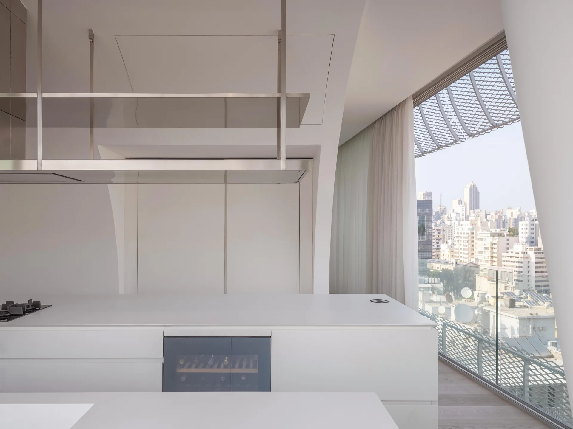 A minimalist white kitchen island with an integrated wine cooler and overhead stainless steel hanging shelves near a window with a city view.