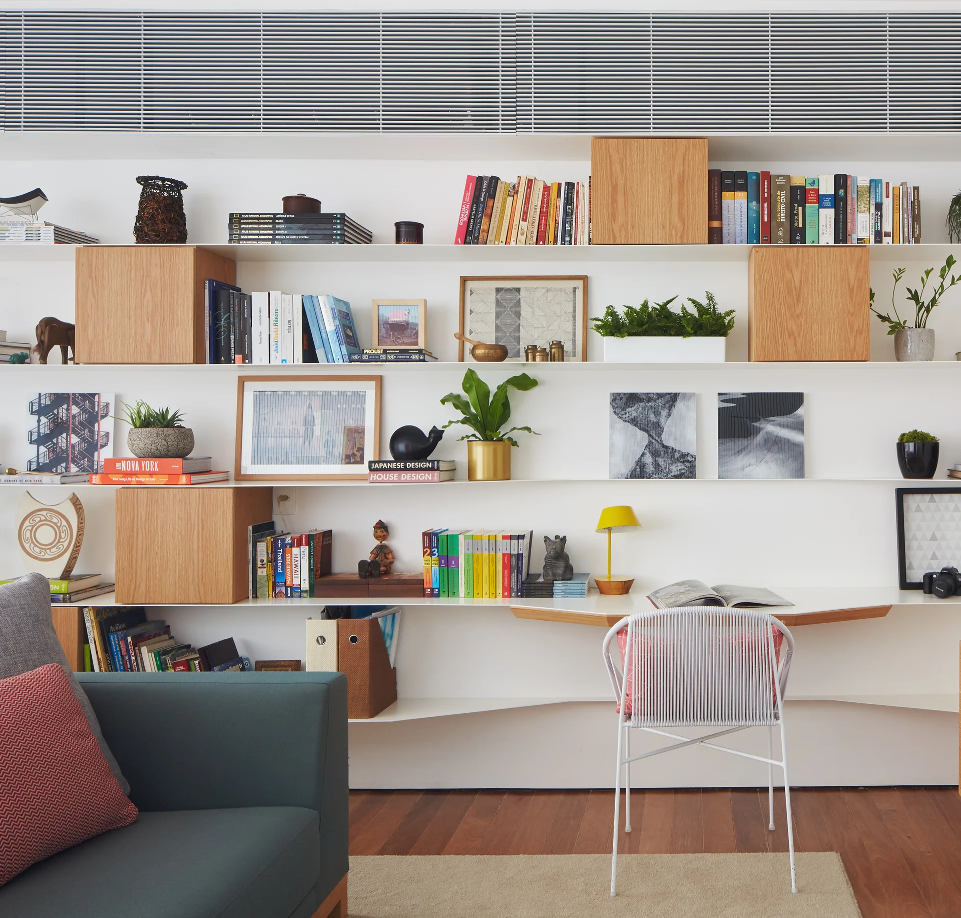 A large white wall-mounted shelving unit features integrated wooden boxes, decorative books, potted plants, and an angled desk with a white wire chair.