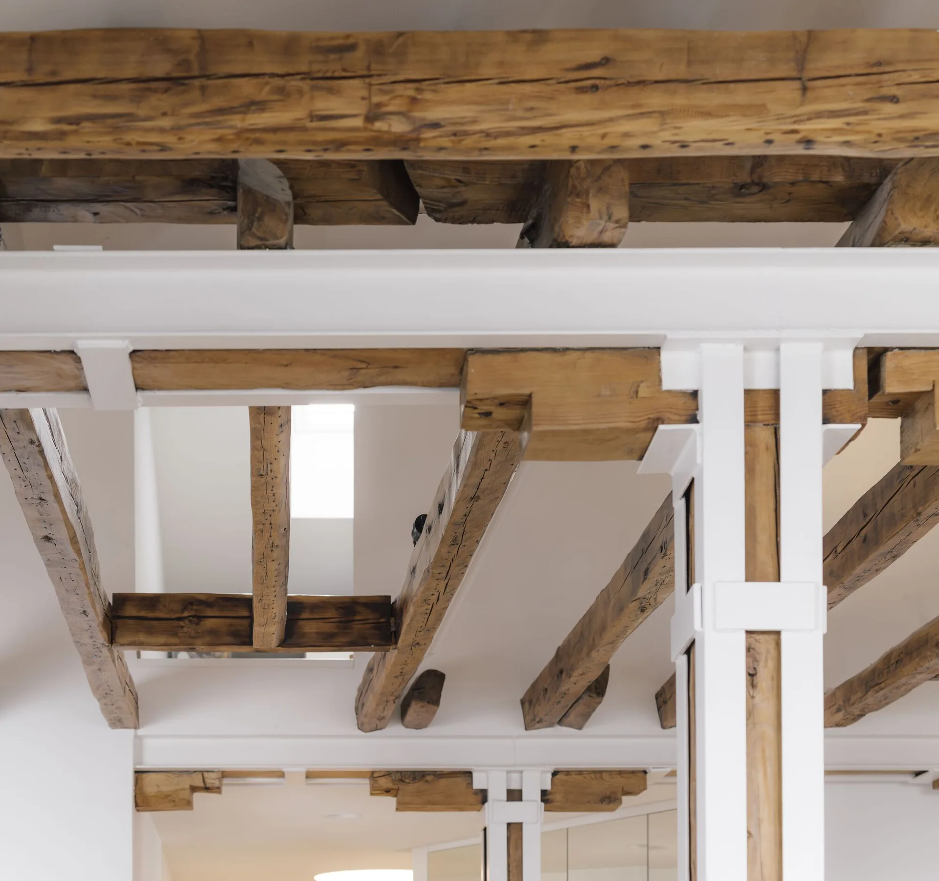 Exposed rustic timber ceiling beams are supported by crisp white painted metal brackets and columns in a minimalist interior.