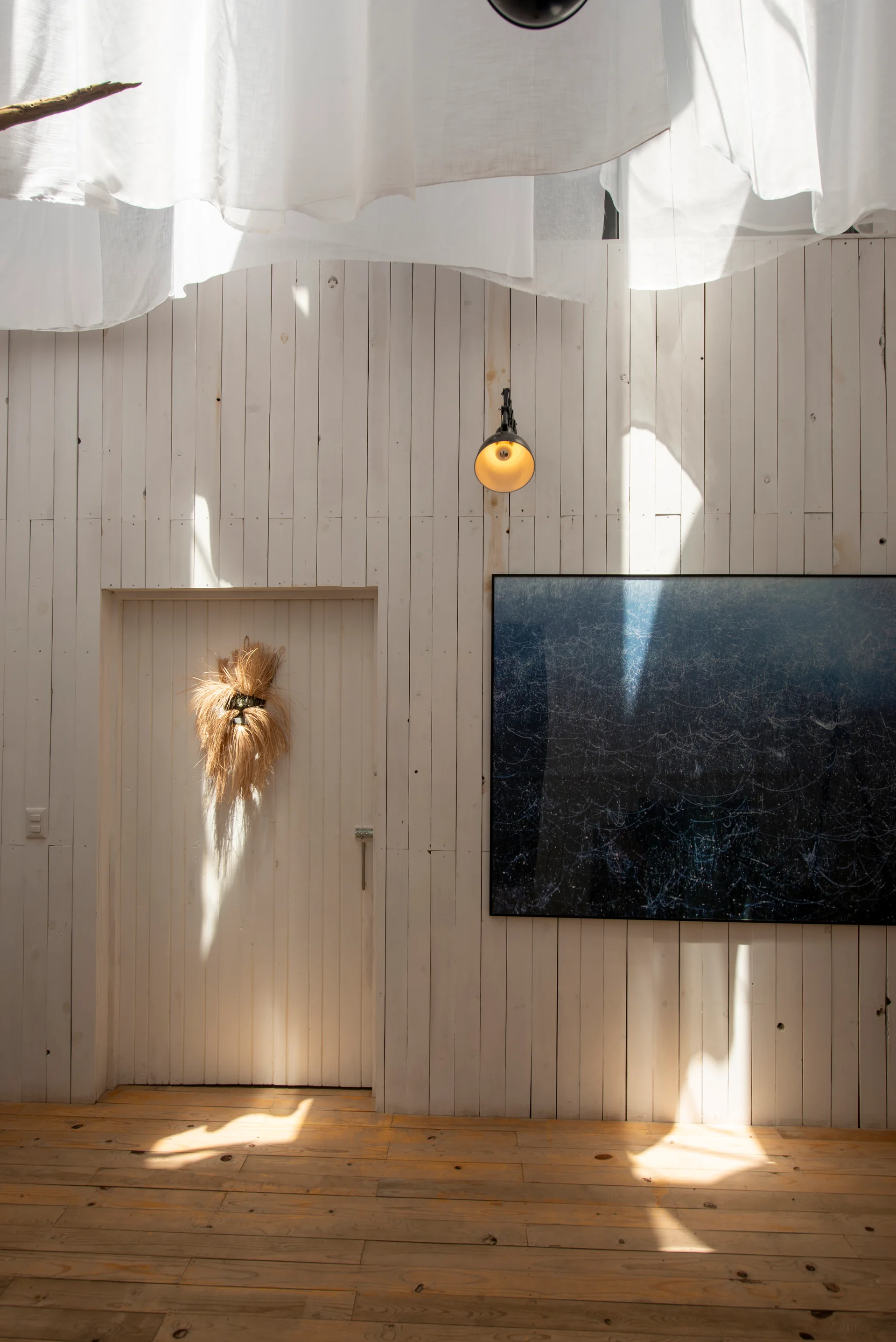 A bright interior features vertical white wood wall paneling, an integrated door with a straw wreath, a large dark framed print, and draped ceiling fabric.