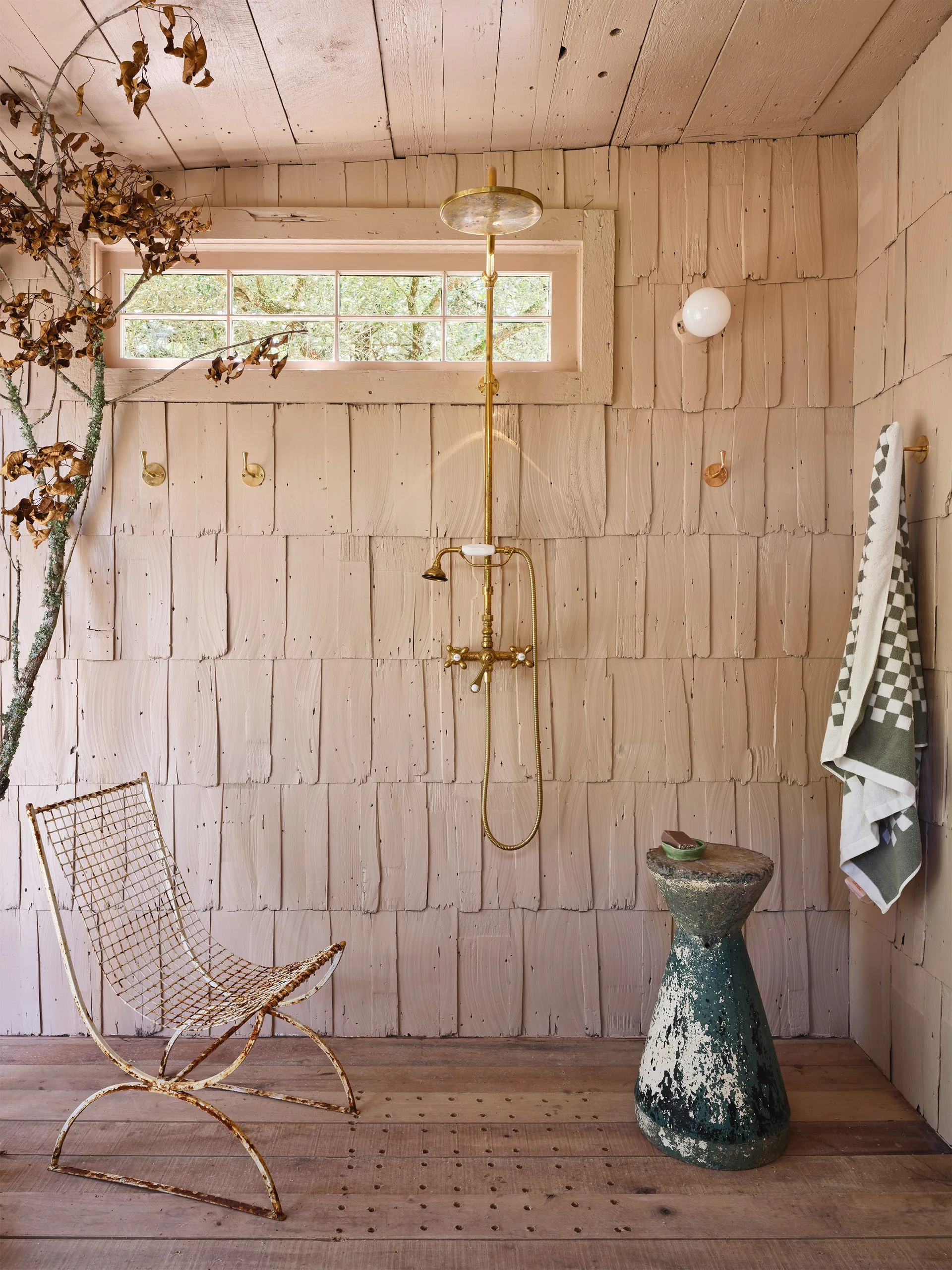A rustic bathroom features pink-painted wood shingle walls, an exposed brass shower fixture, a weathered metal chair, and a distressed green stool.