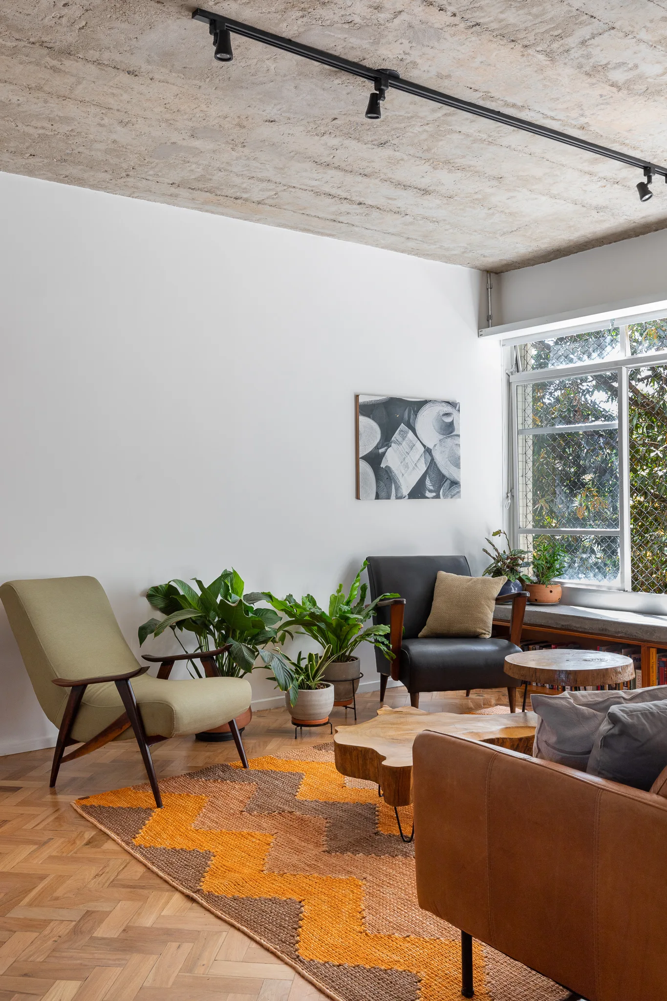 A living room featuring an exposed concrete ceiling, herringbone wood parquet floors, and mid-century modern armchairs with natural wood coffee tables.