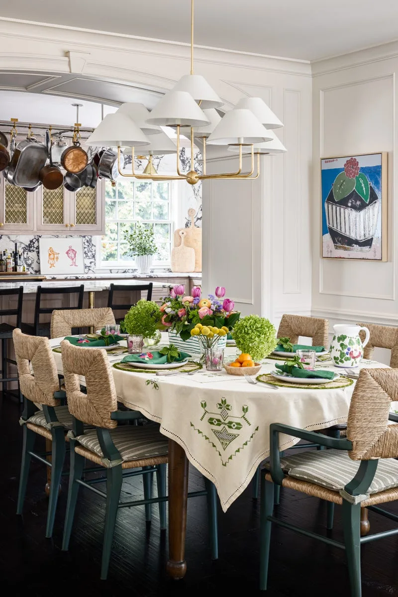 A dining room features a wooden table with an embroidered tablecloth, green-painted woven chairs, and a white multi-shade chandelier.