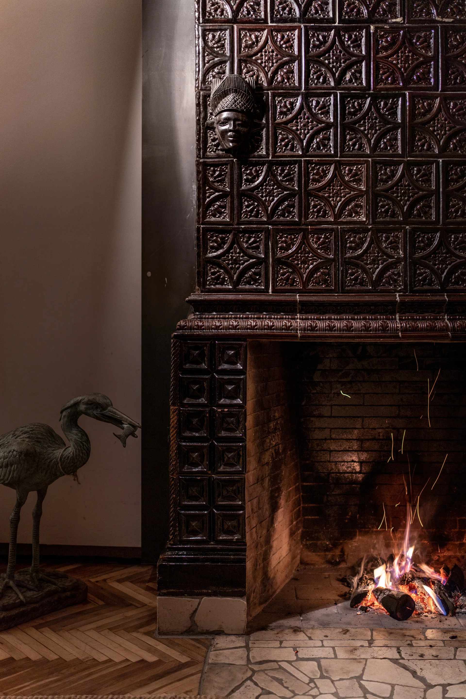 An ornate fireplace built with glossy dark brown patterned ceramic tiles stands next to a bronze bird statue on herringbone wood floors.