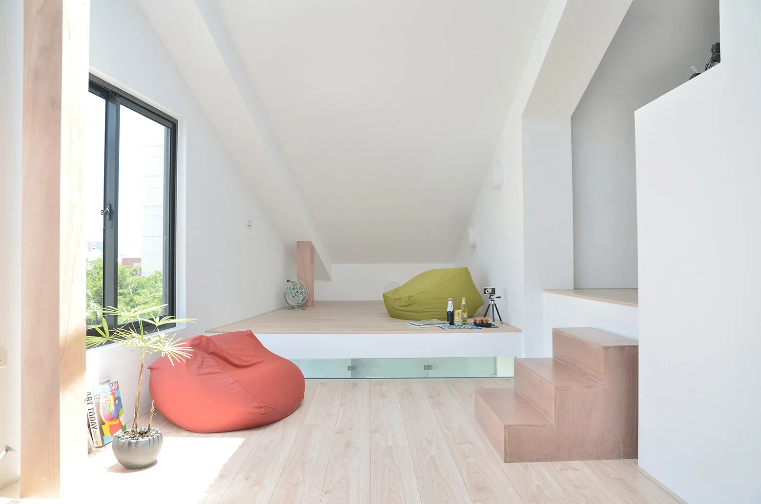 A minimalist split-level living space with white walls, a raised wooden platform, wooden stairs, and red and green bean bag chairs.