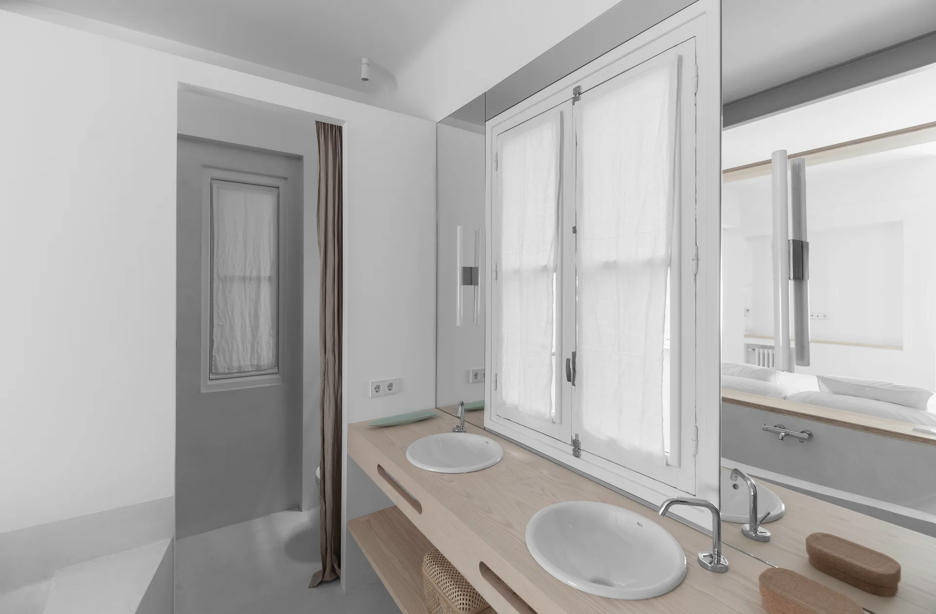 A minimalist bathroom features a long light wood vanity with dual white sinks, a large mirror integrated around a double casement window, and a woven basket.