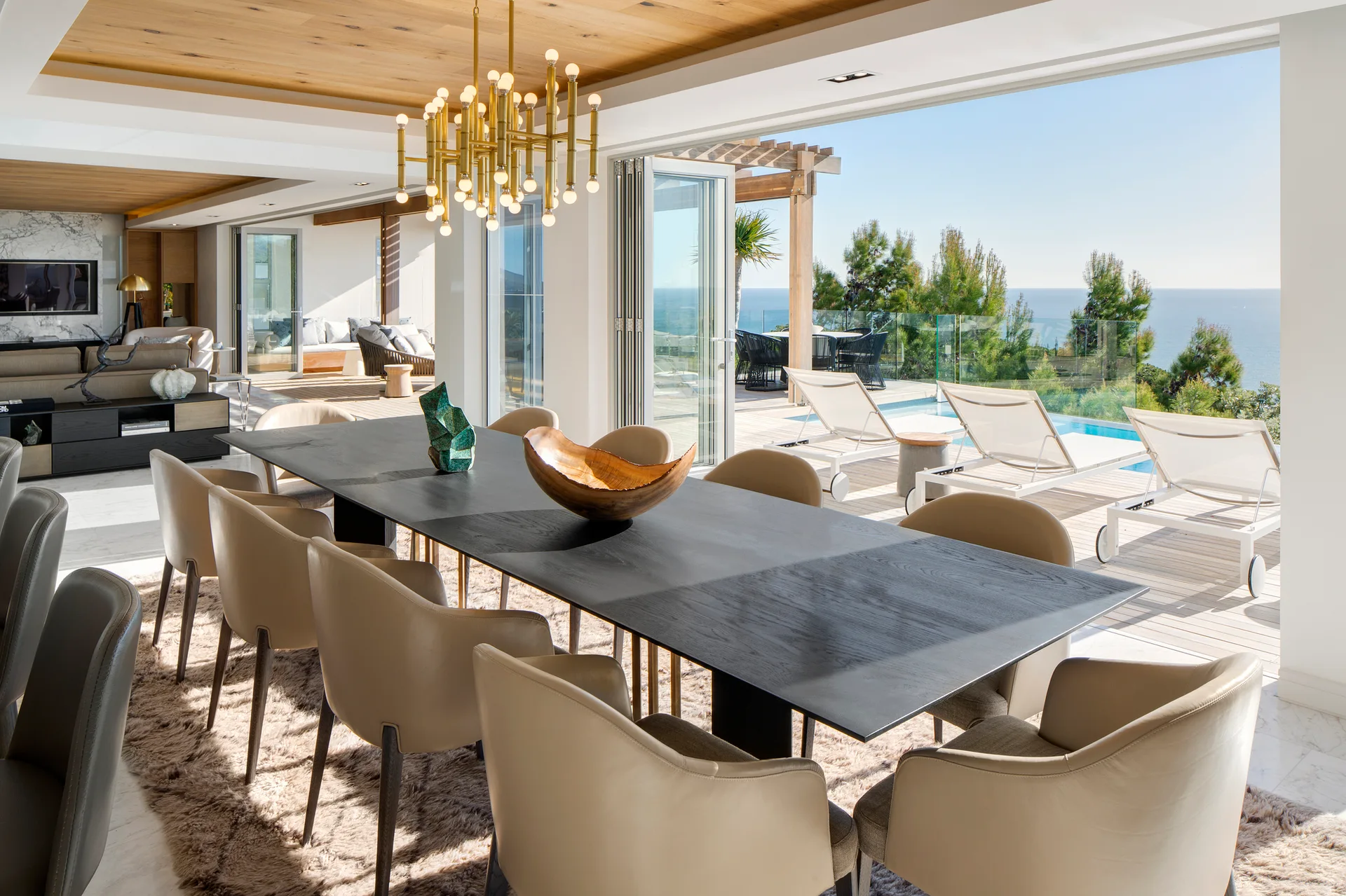 A contemporary dining room features a black table with tan chairs, a gold chandelier, and open bi-fold doors leading to a pool deck.