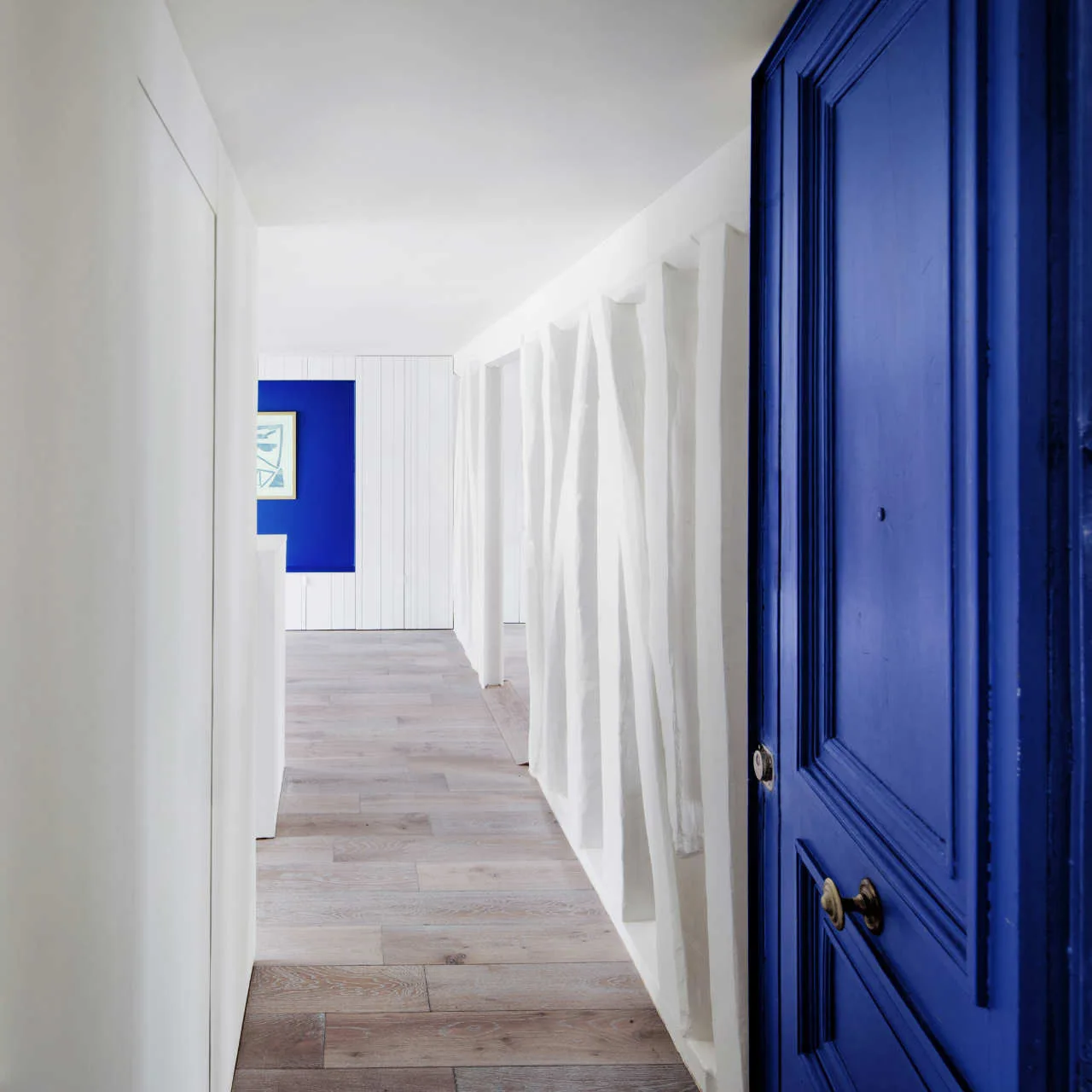 A long white corridor features a prominent cobalt blue door in the foreground, vertical wall panels, and light wood flooring.