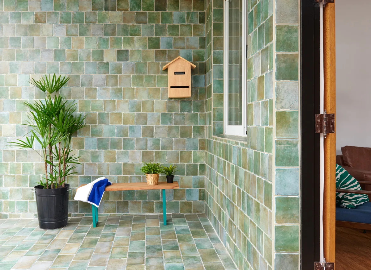 An outdoor patio features green glazed square tiles on the walls and floor, styled with a wooden bench, potted plants, and a wooden mailbox.