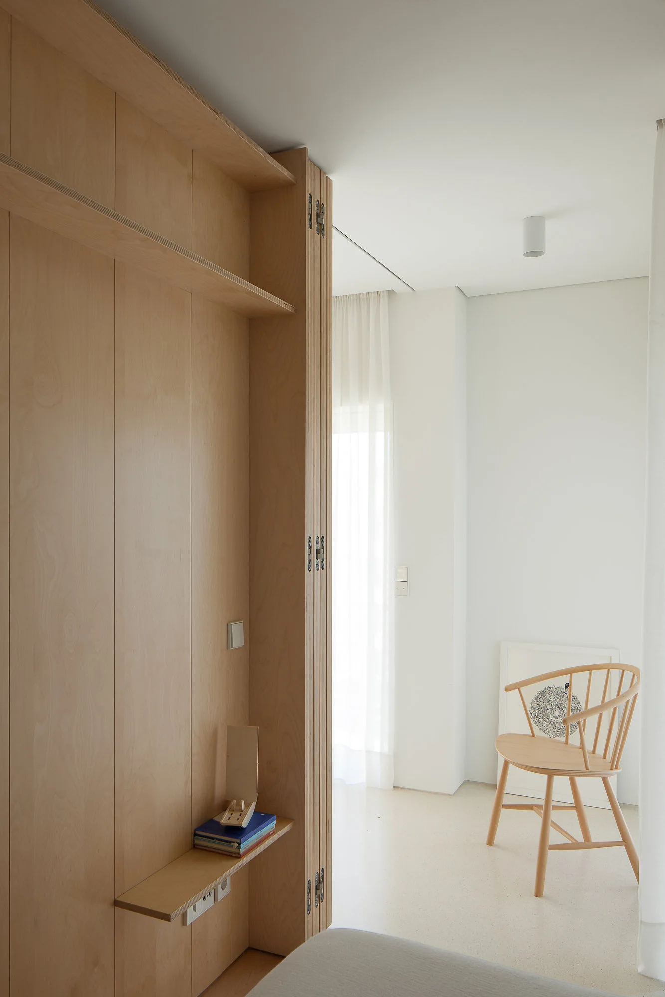 A full-height light wood storage cabinet stands next to a white wall with a wooden spindle back chair and sheer curtains in the background.