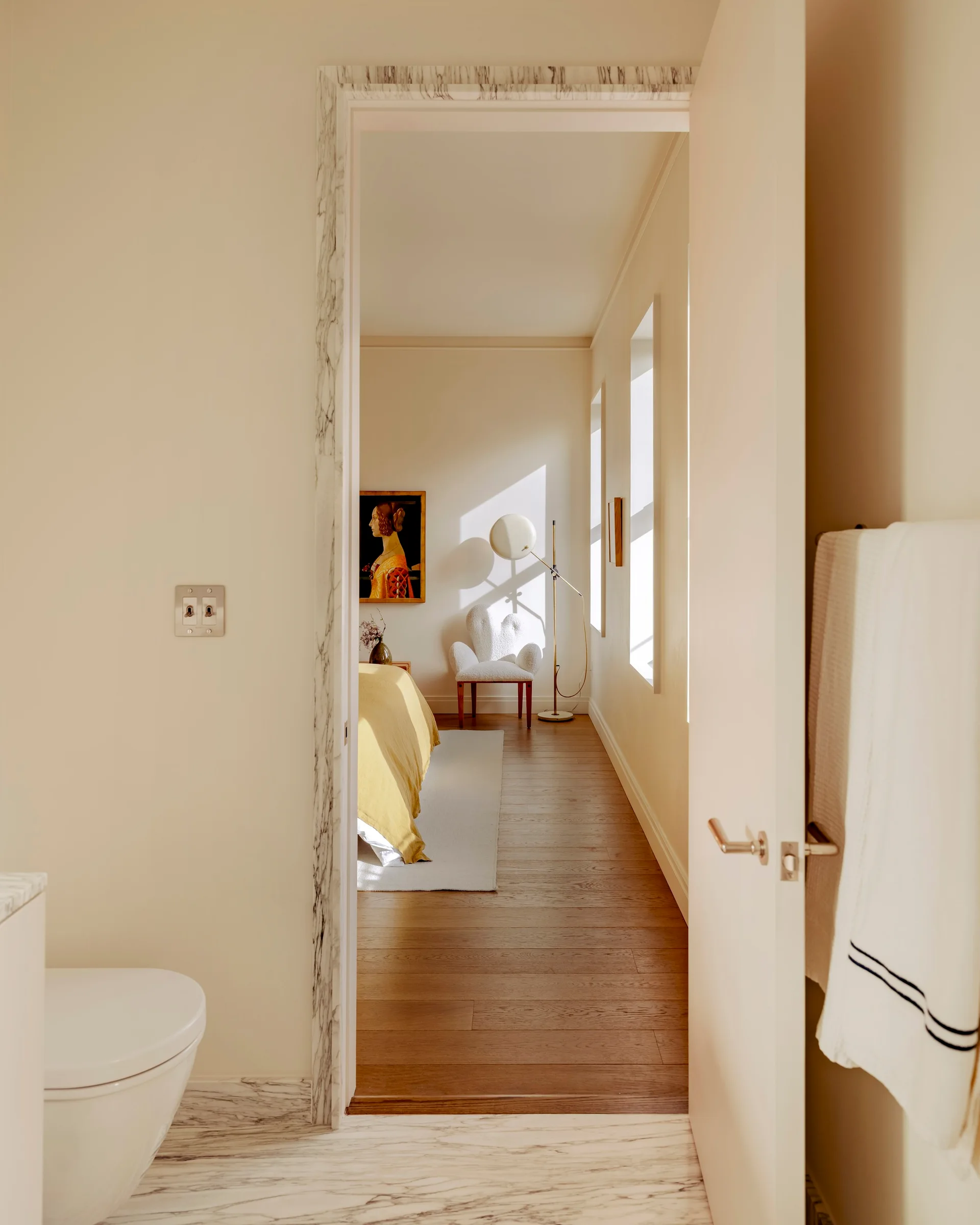 A view through a marble-trimmed doorway reveals a warm minimalist bedroom featuring a bed with yellow bedding, a textured armchair, and wood floors.