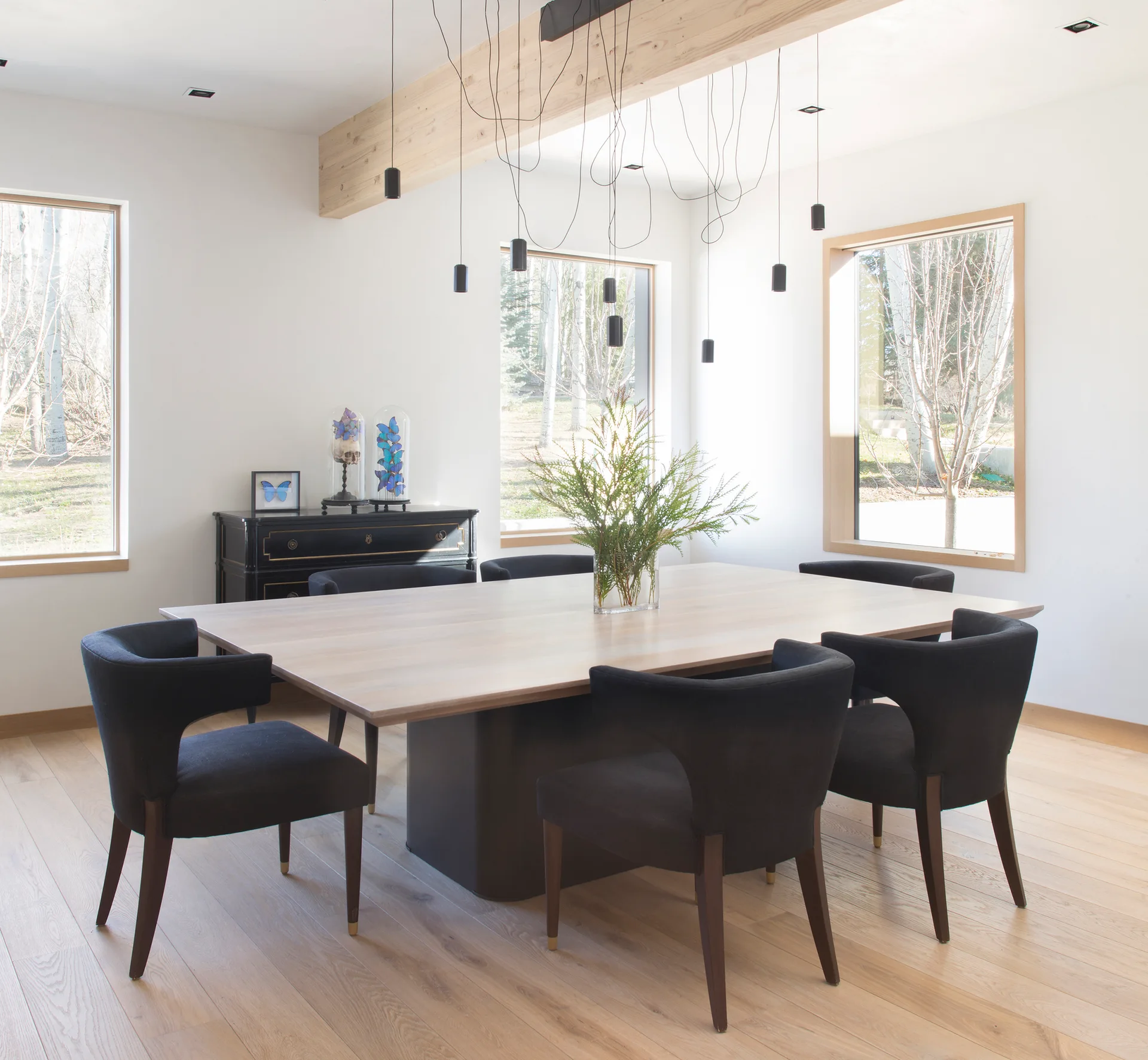 A modern dining room features a light wood table surrounded by black upholstered chairs under a cluster of slim pendant lights and an exposed ceiling beam.