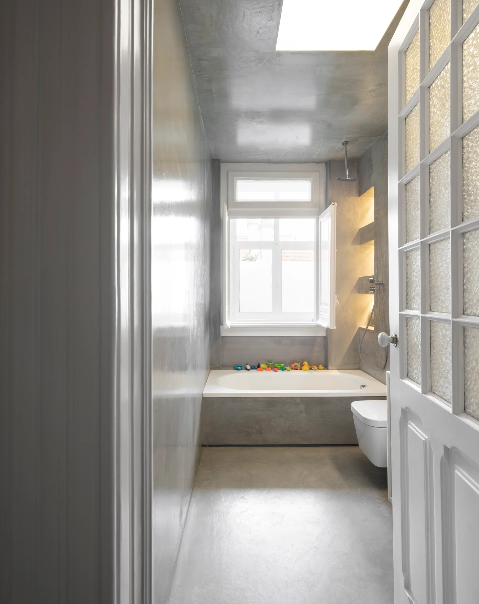 A narrow grey concrete bathroom features a white built-in bathtub under a window, a skylight, and recessed wall niches with warm lighting.