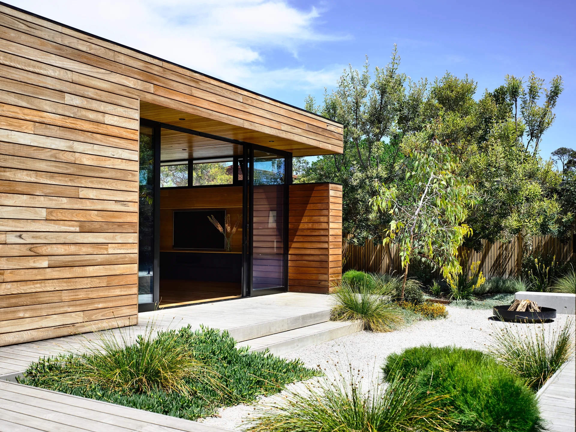 A modern wood-clad pavilion with wide sliding glass doors opening onto a light timber deck, facing a landscaped gravel garden with native plants.