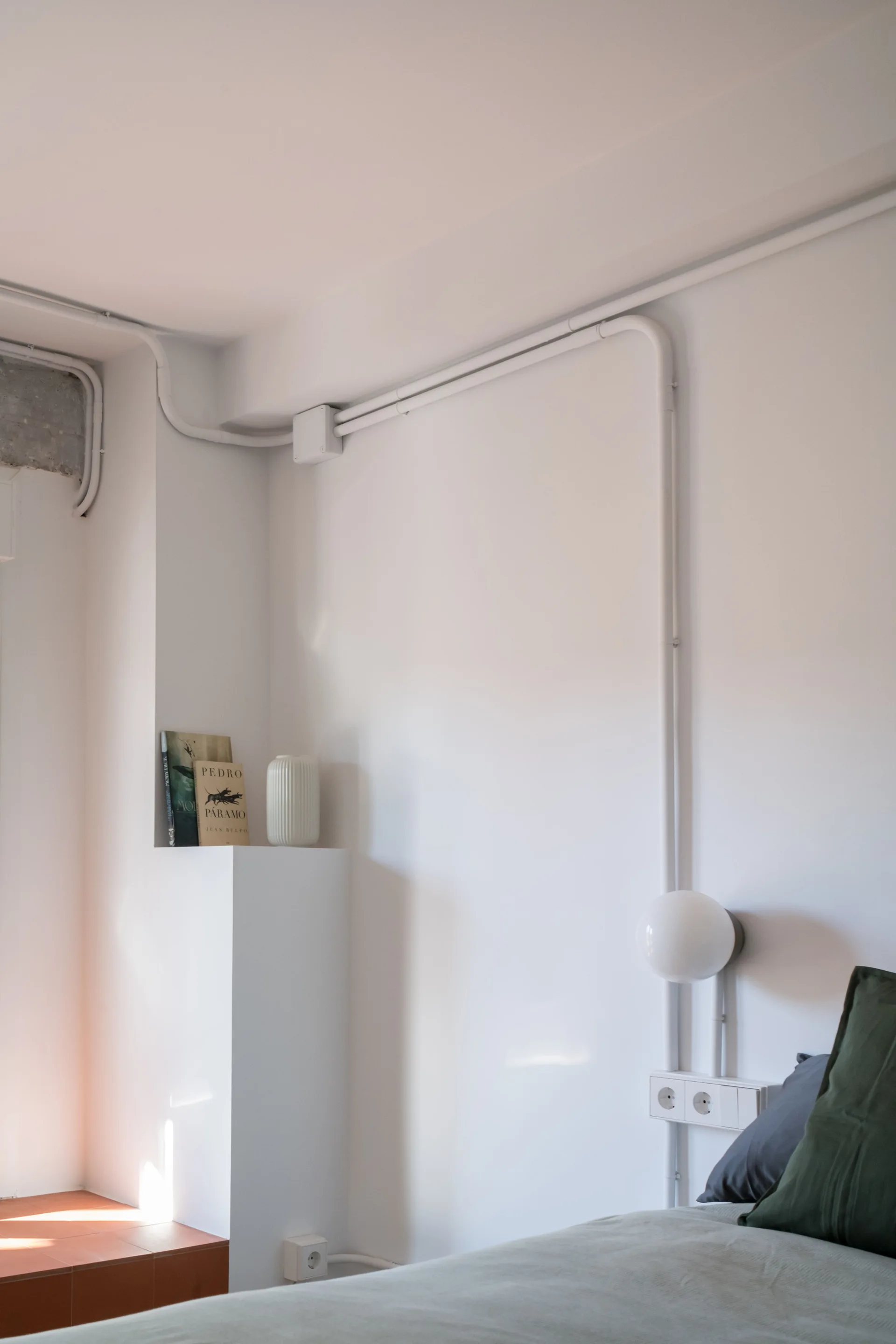 A minimalist bedroom corner shows a bed with green pillows next to a white wall with exposed painted conduit, a globe sconce, and a built-in ledge.