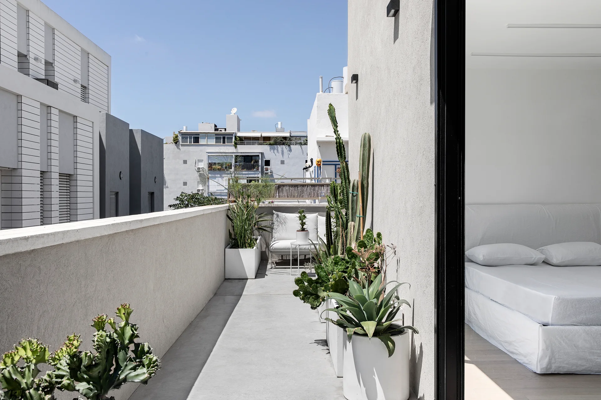 A minimalist white balcony features large potted cacti and a lounge chair next to an open sliding door revealing an all-white bed.