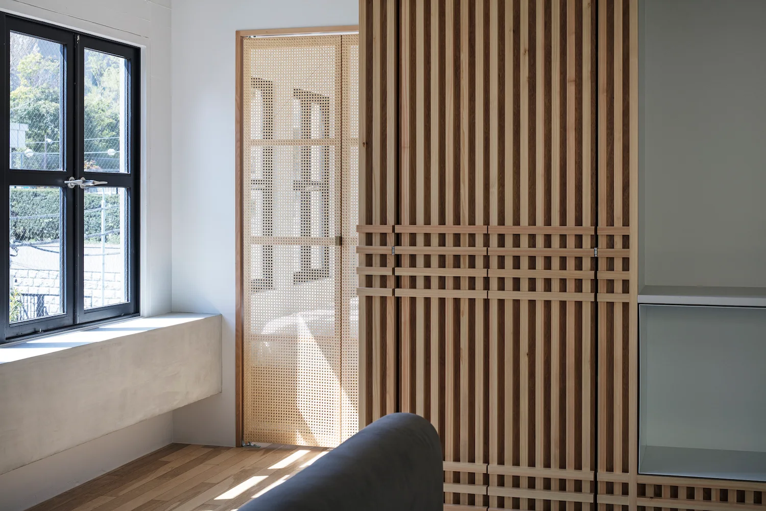 An interior space featuring a vertical slatted wood partition wall, a folding door with cane webbing panels, and a black-framed window.