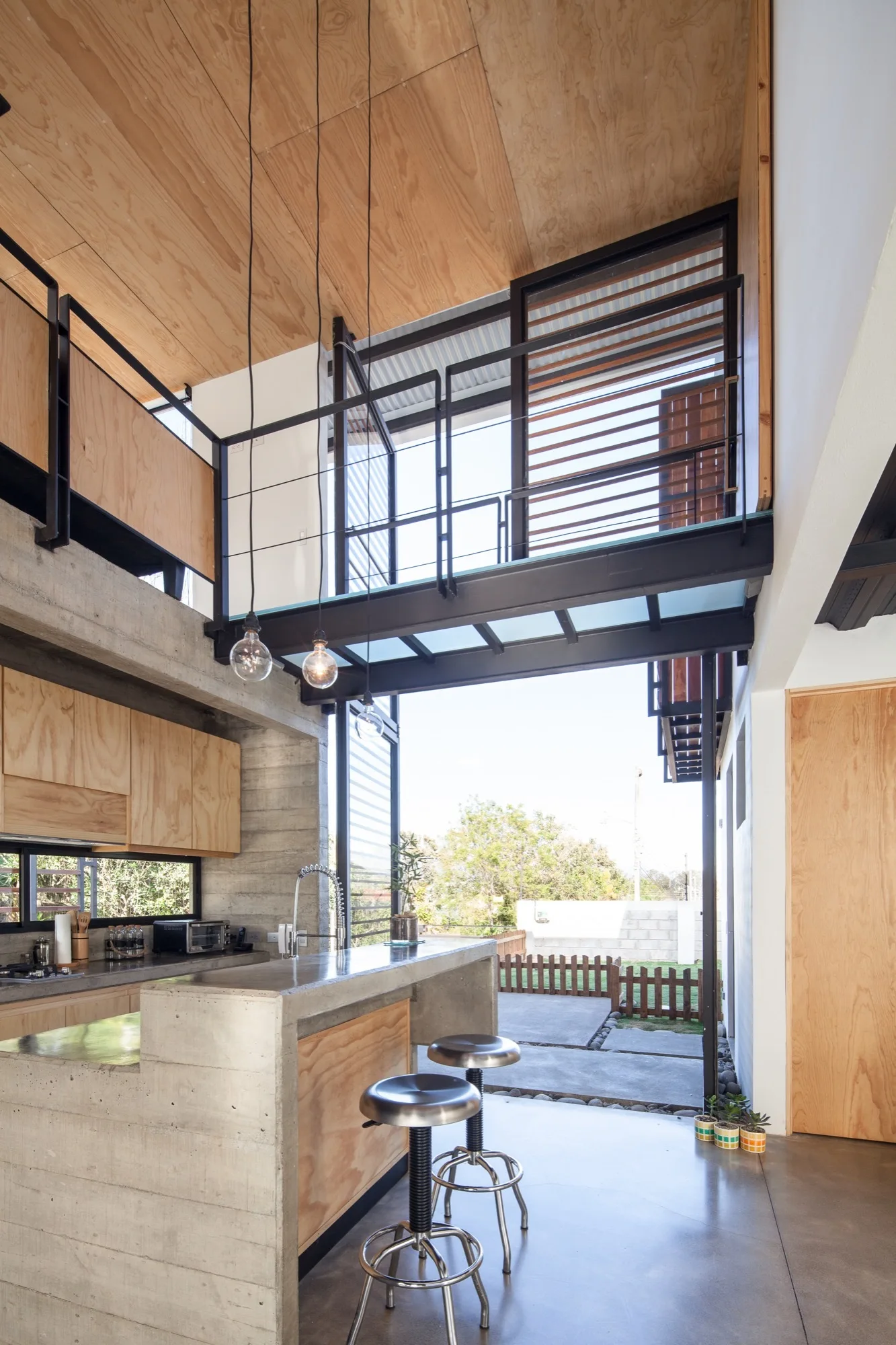 An industrial kitchen with a concrete island, light wood cabinets, and metal bar stools below an open mezzanine with black metal railings.