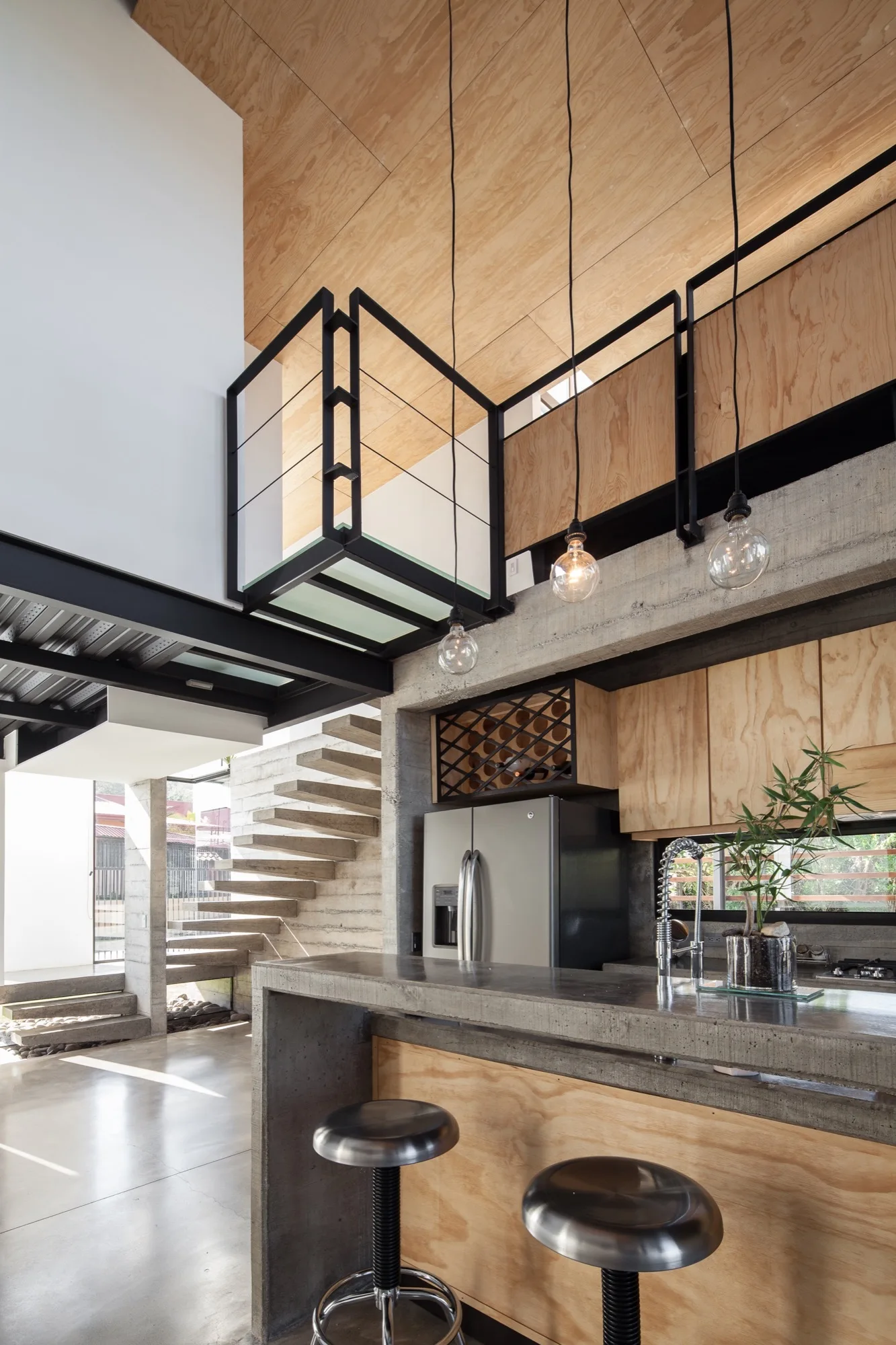 An industrial kitchen featuring a concrete island with metal stools, natural plywood cabinetry, and a floating concrete staircase in the background.