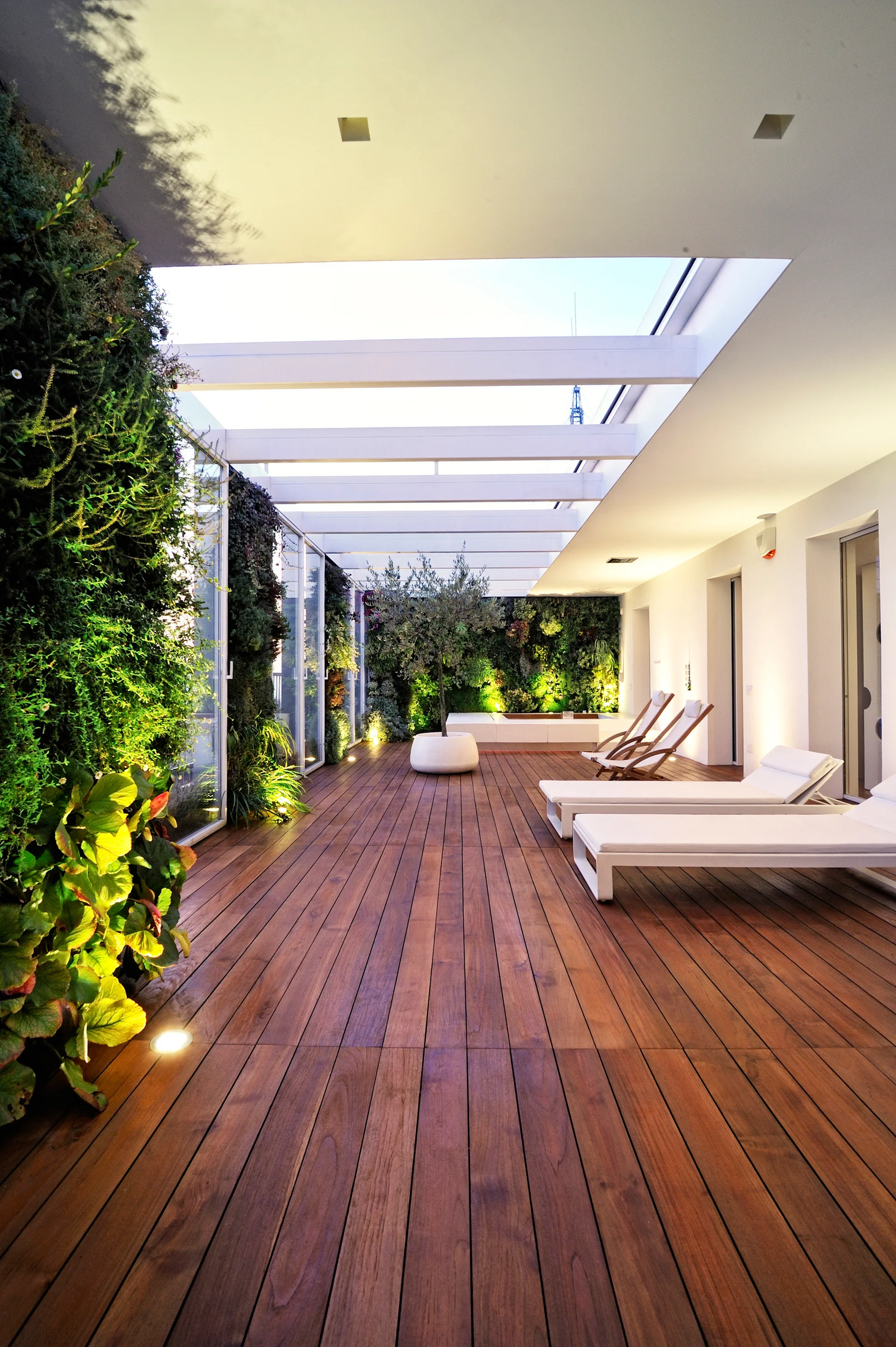 A modern outdoor terrace featuring wooden plank decking, white sun loungers, and lush vertical gardens under an open white pergola.