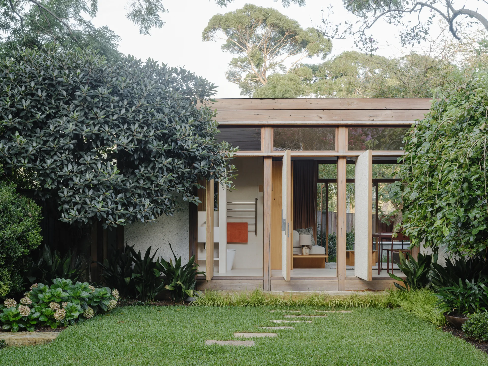 A modern timber-framed pavilion with large open doors facing a green lawn with a stone stepping path and lush garden foliage.