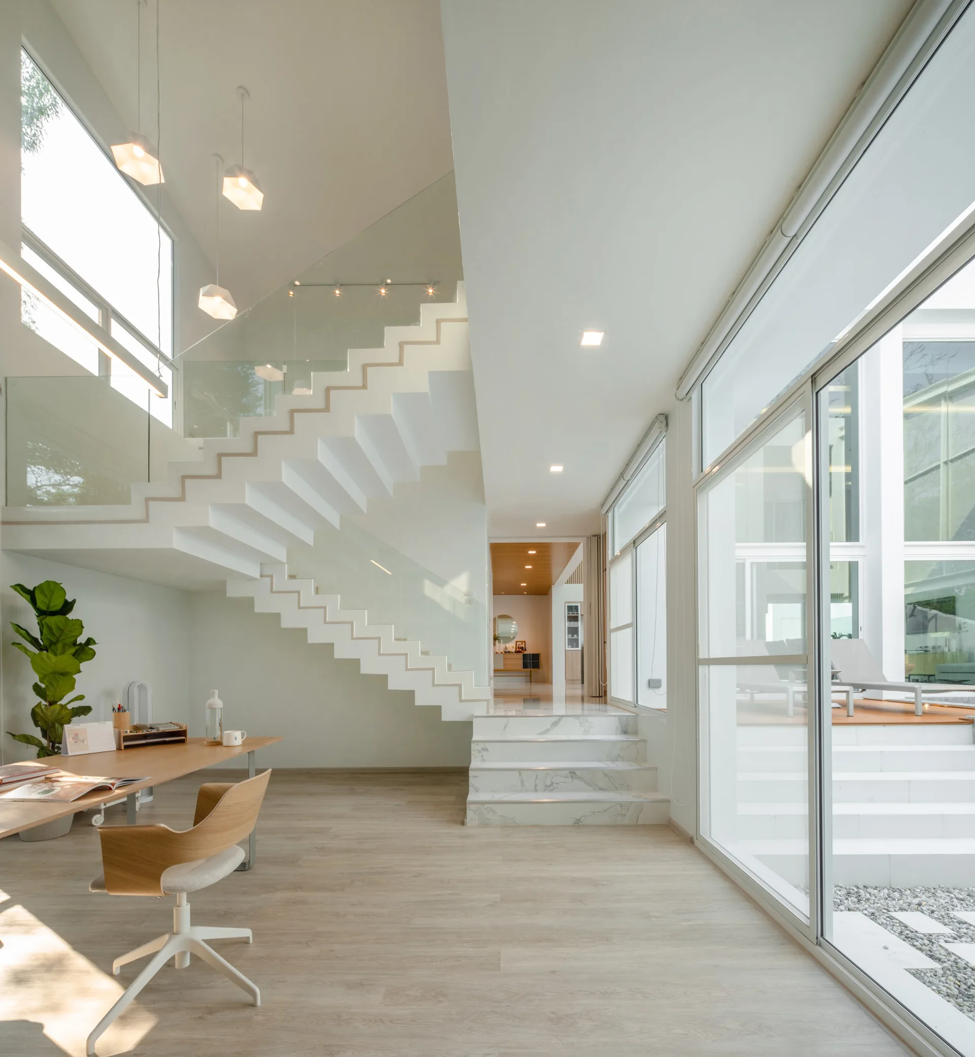 A spacious modern room features a white floating staircase with glass railings, a light wood desk with a chair, and large sliding glass doors.