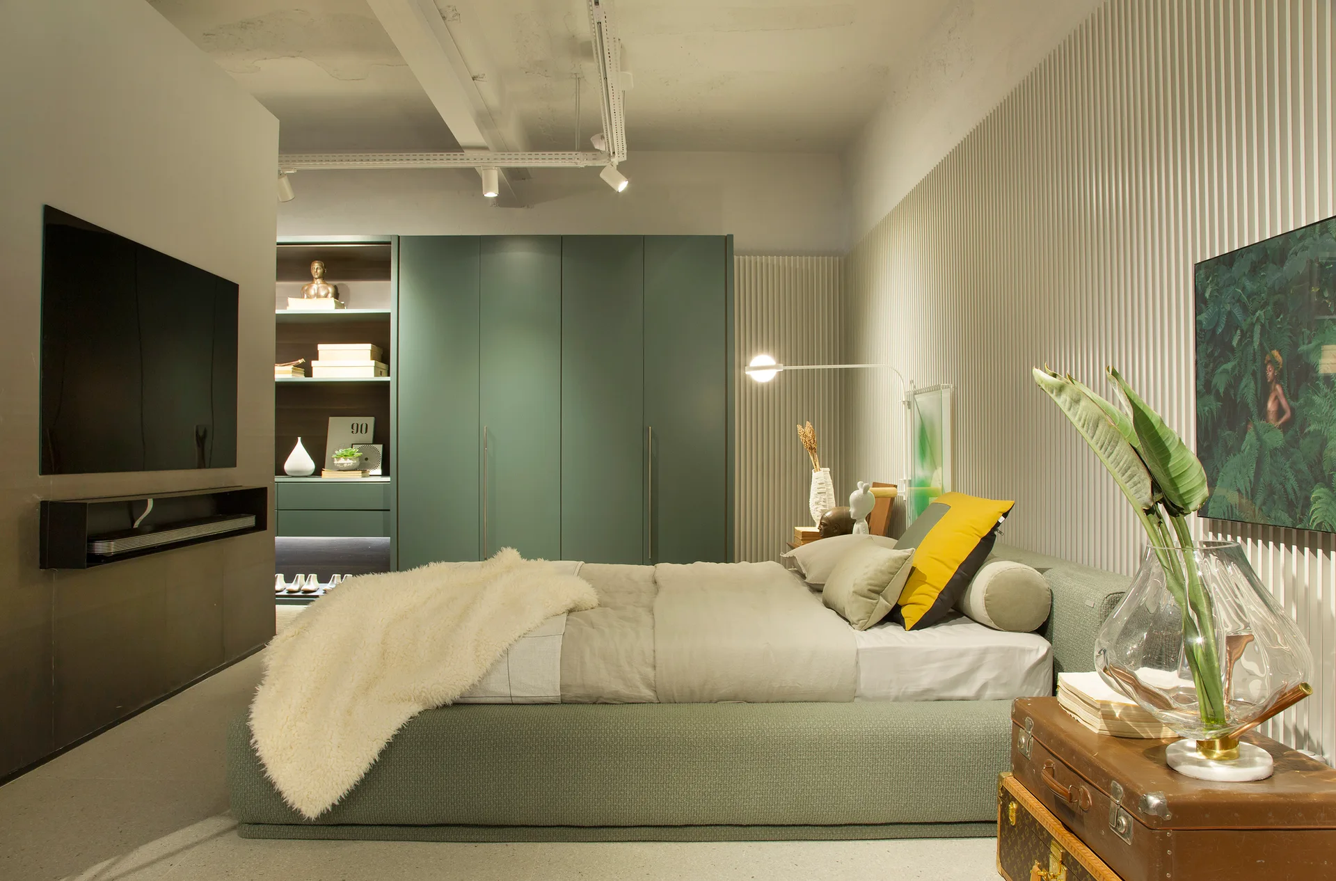 A low sage green bed is set against a vertical slatted wall next to built-in green wardrobes, illuminated by ceiling track lights.