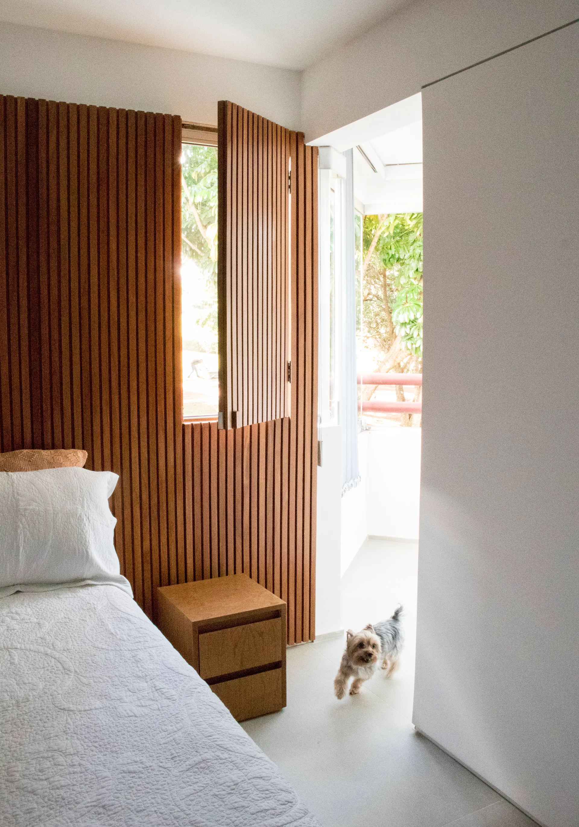 A modern bedroom features a white bed next to a warm wood slatted wall with an integrated shutter window opening to natural light.