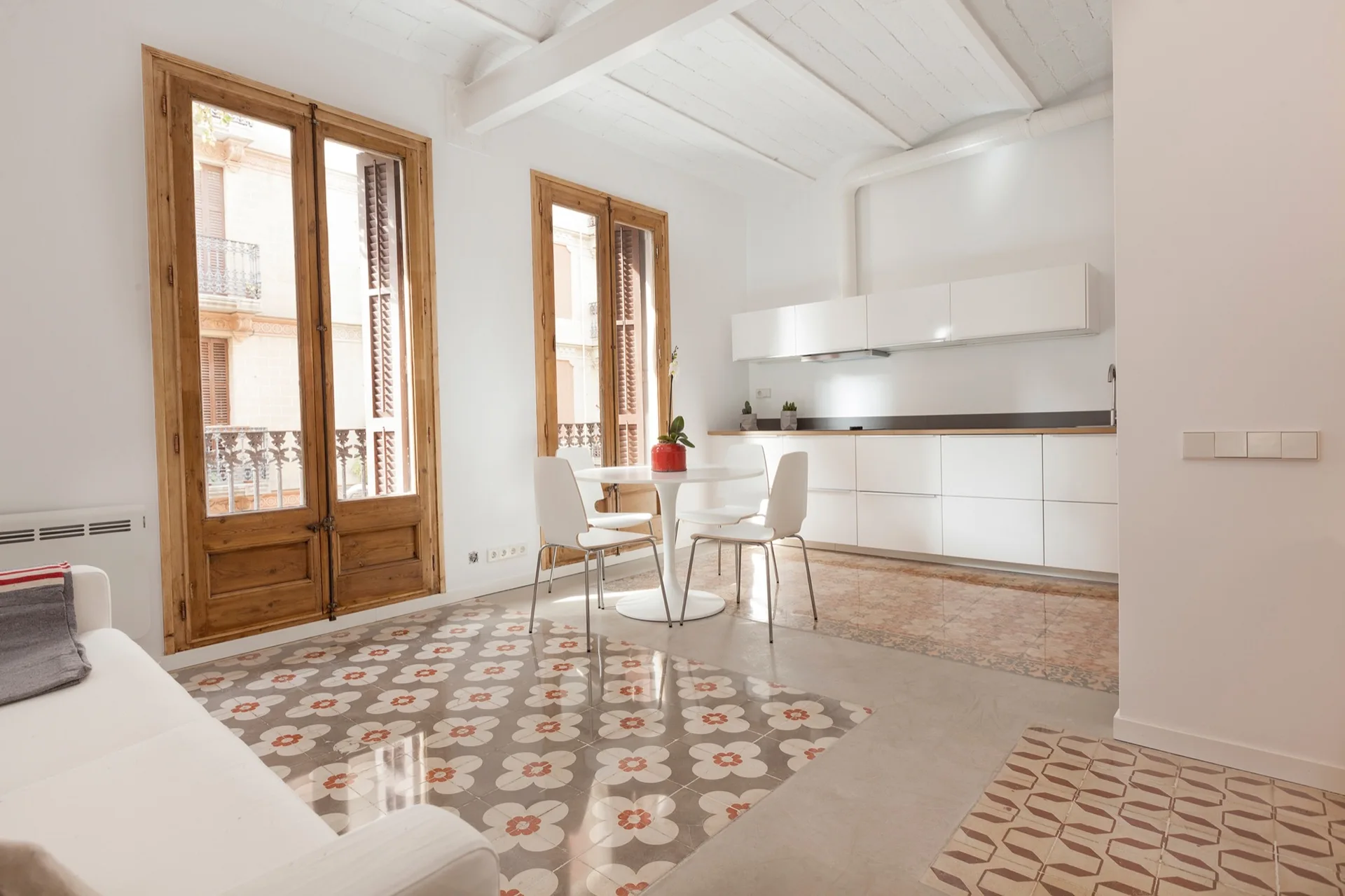 A bright kitchen and dining area features white minimalist cabinets, a round dining table, and tall wood-framed balcony doors above patterned tile flooring.