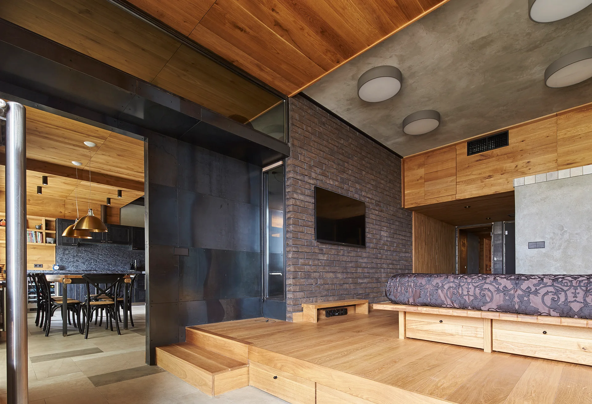 A modern bedroom with an elevated wooden platform bed against a dark brick wall next to a metallic partition and concrete ceiling.