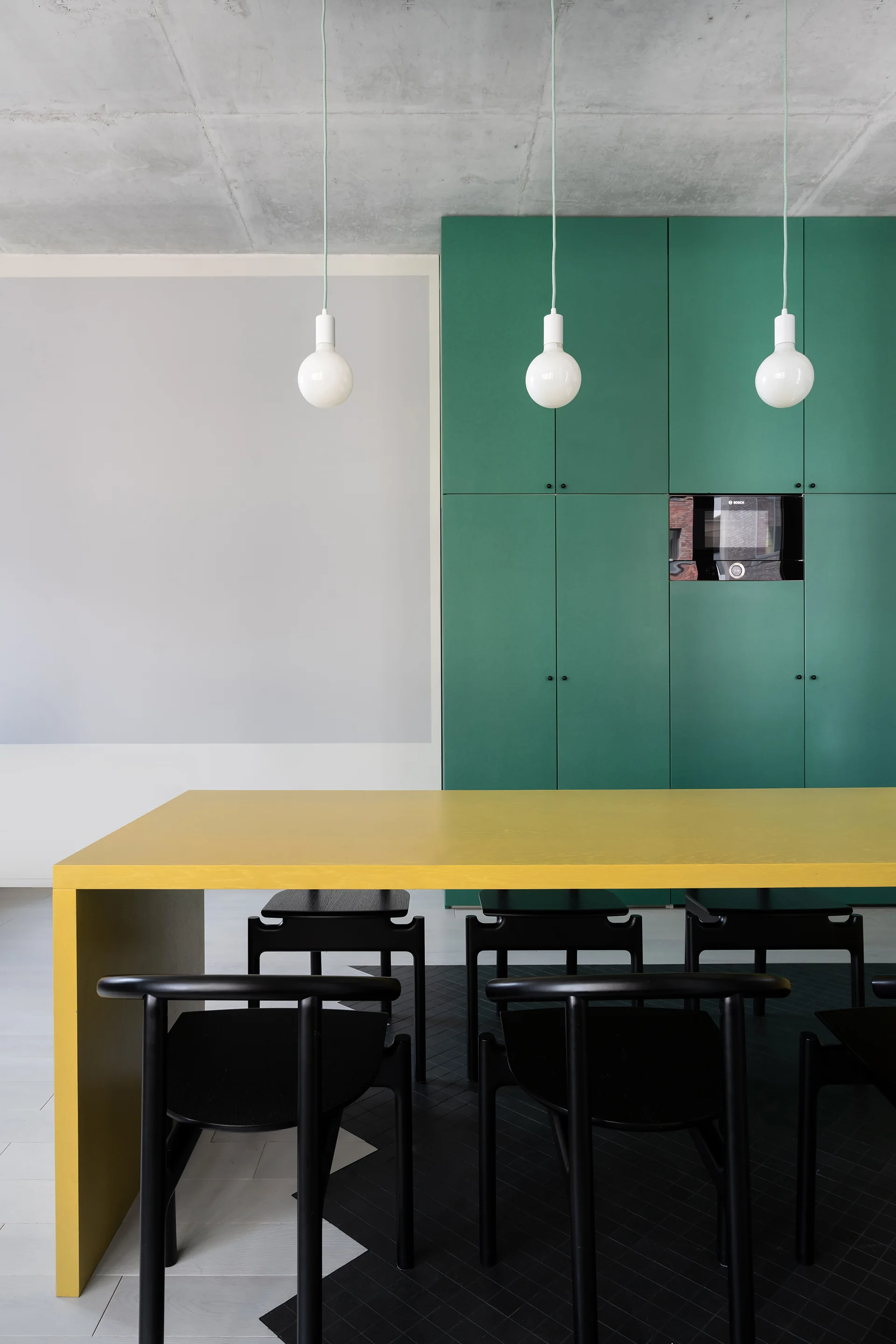A bright yellow dining table and black chairs are set against matte green cabinetry and an exposed concrete ceiling.
