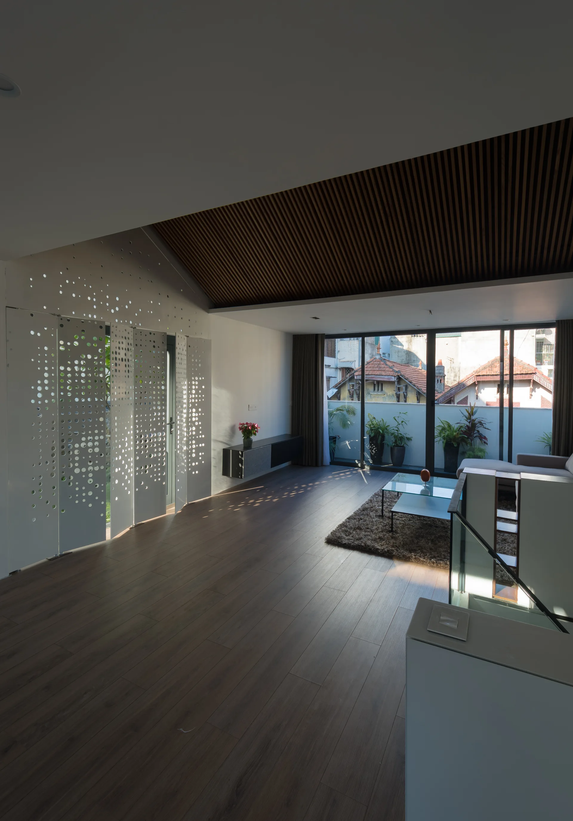 A minimalist living room with wood floors, a slatted wood ceiling, a white perforated metal screen, and glass doors opening to a balcony.