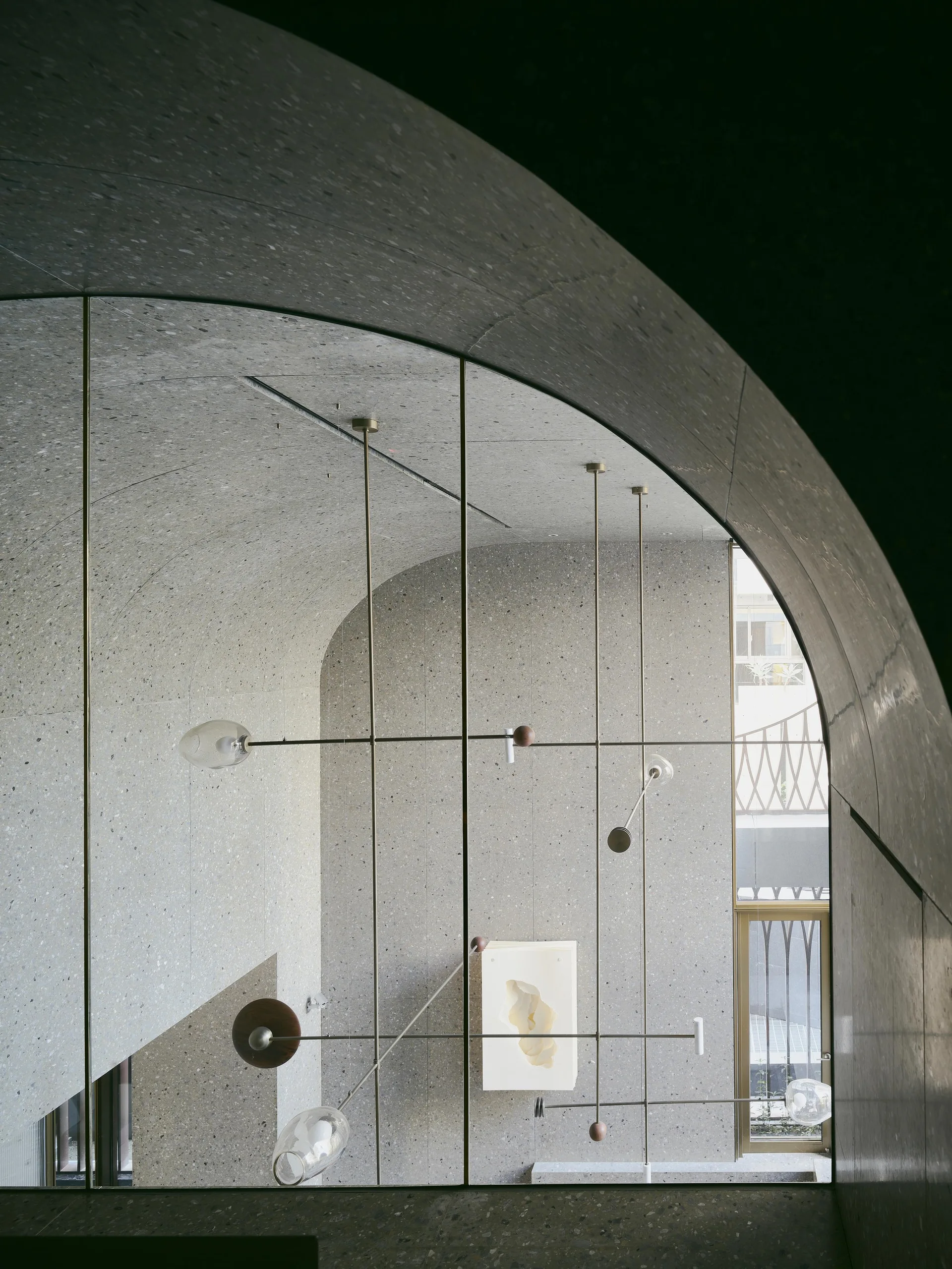 A minimalist architectural interior featuring curved grey terrazzo walls, an arched ceiling, and a slender multi-arm metal pendant light fixture.