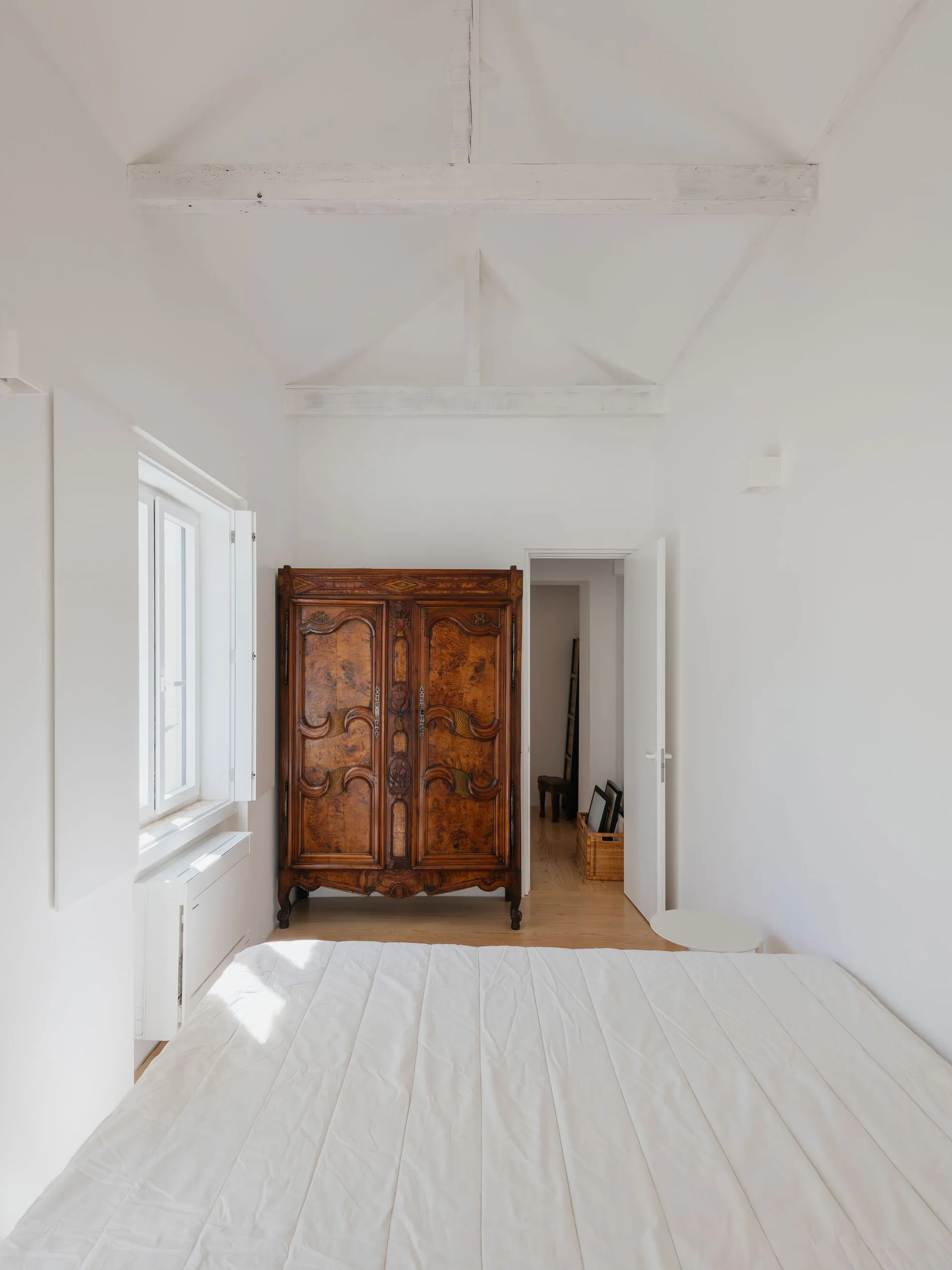 A bright minimalist bedroom features a bed with a white comforter, white vaulted ceilings with beams, and a large antique wooden wardrobe.