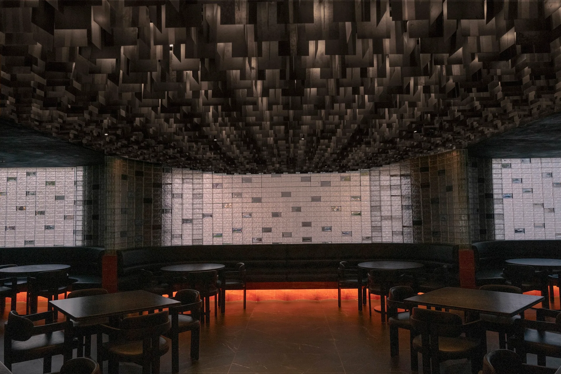 A dark commercial lounge features curved black banquette seating beneath a geometric ceiling grid, set against a glowing glass block wall.