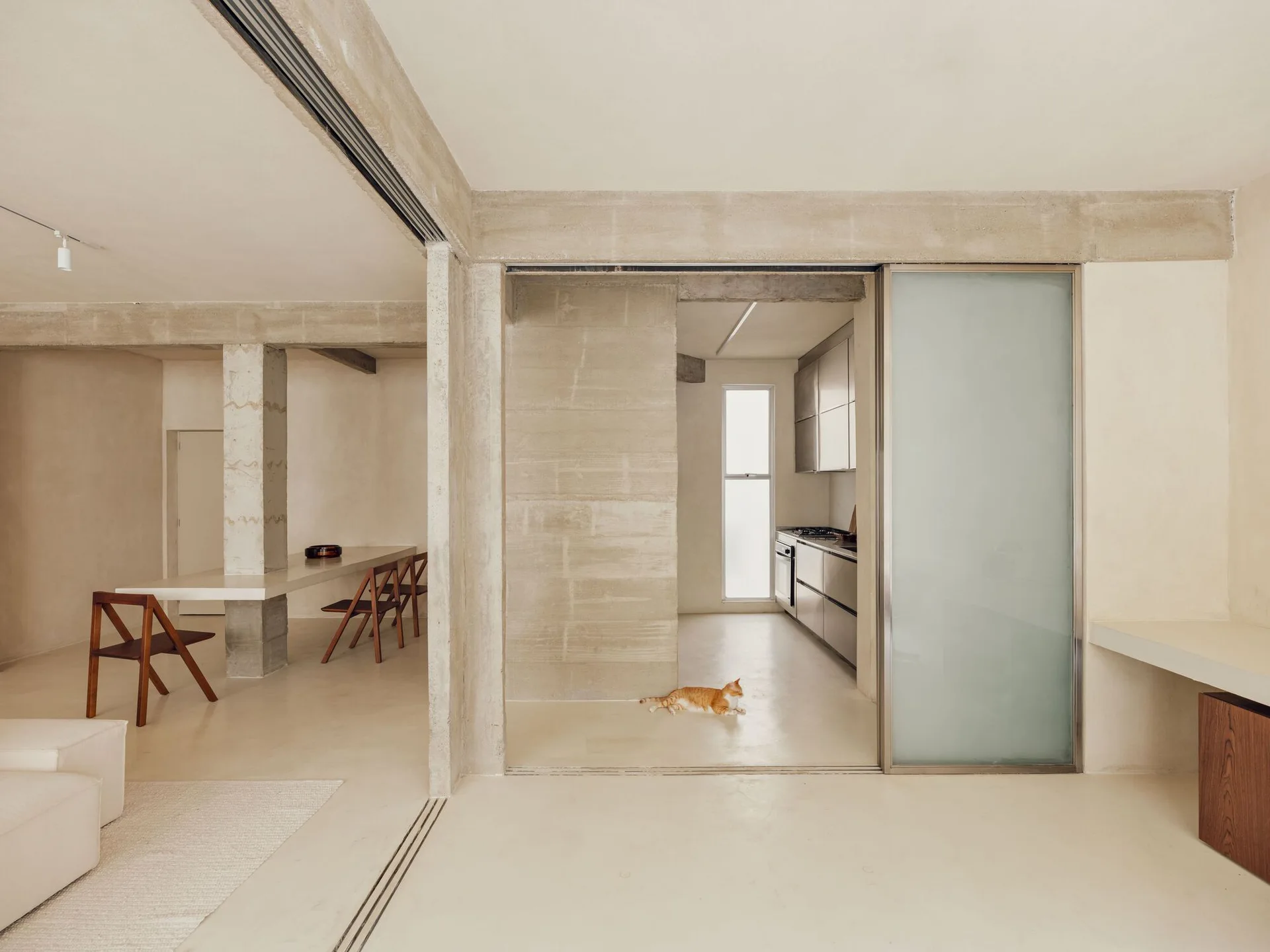 A minimalist open-plan interior features raw concrete walls, a long white dining table integrated with a structural column, and a sliding glass partition.