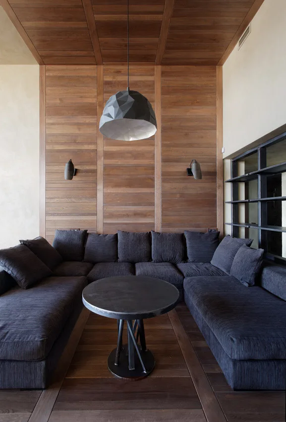 A modern lounge featuring a dark grey sectional sofa, a round metal coffee table, and warm wood paneling on the wall and ceiling.