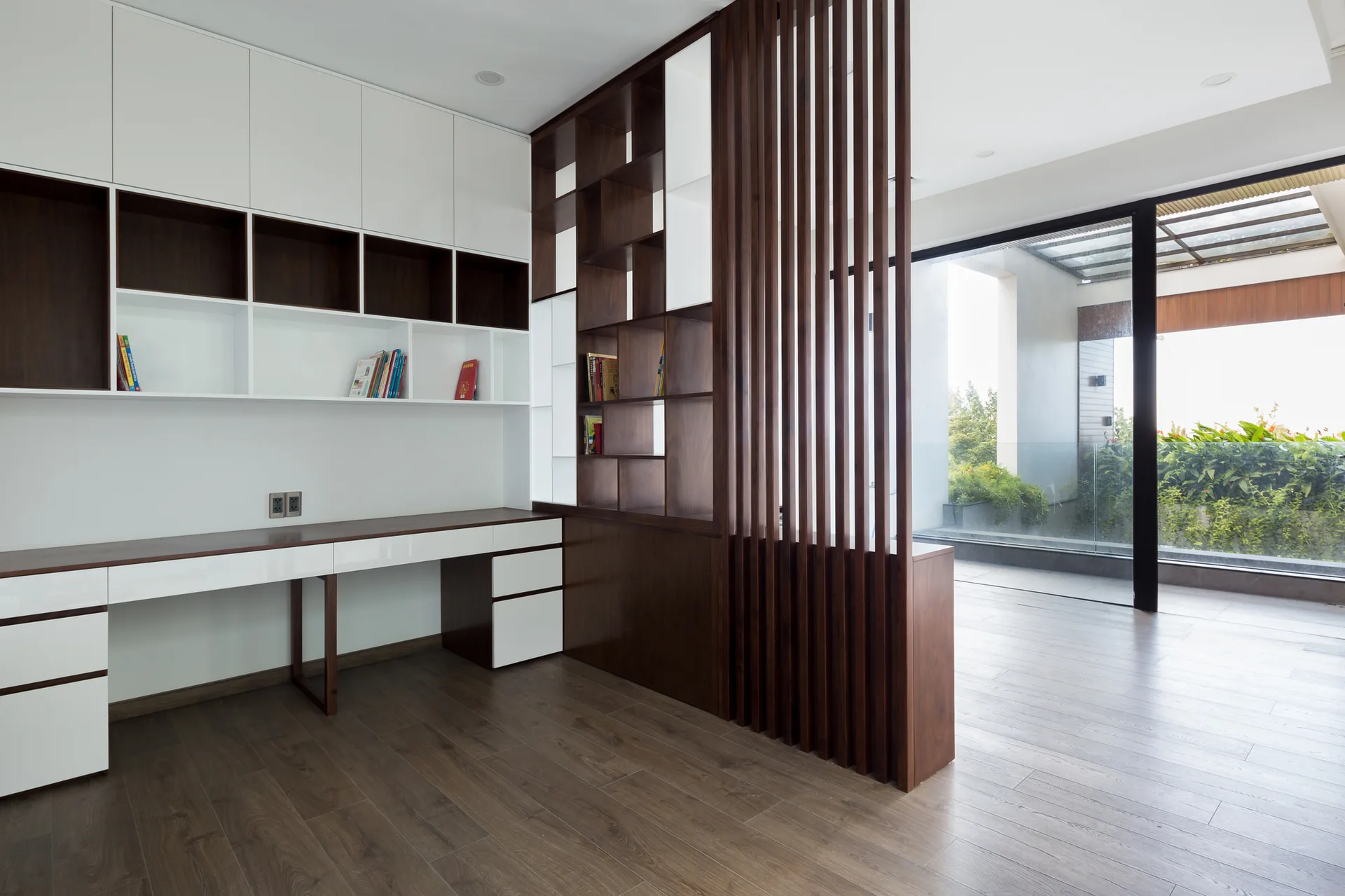A modern home office features a built-in white and wood desk, wall-mounted shelving, and a dark wood vertical slat room divider next to glass sliding doors.