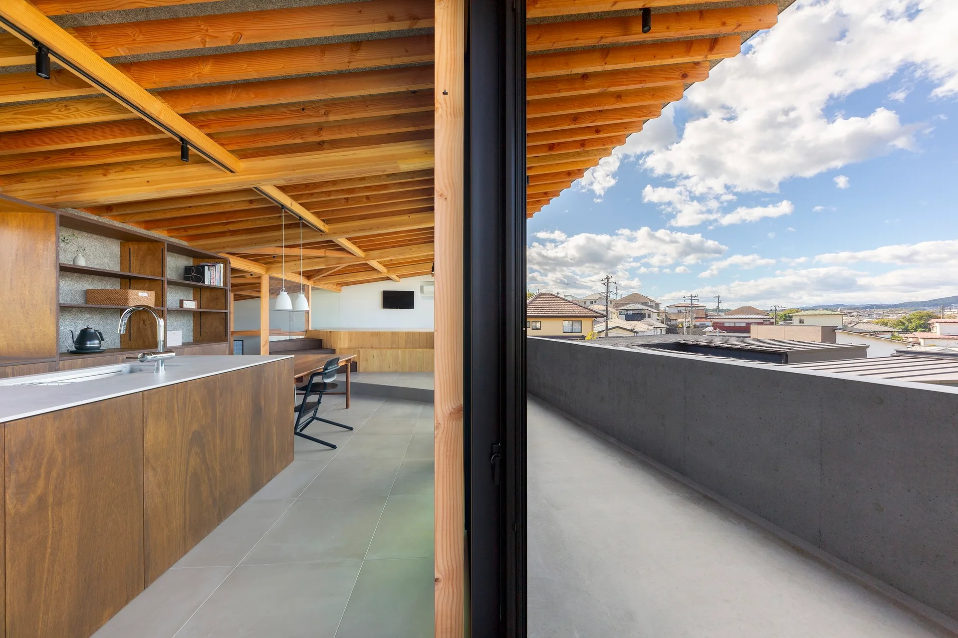 A modern wooden kitchen with an island and exposed ceiling beams opens onto a concrete balcony under a cloudy blue sky.