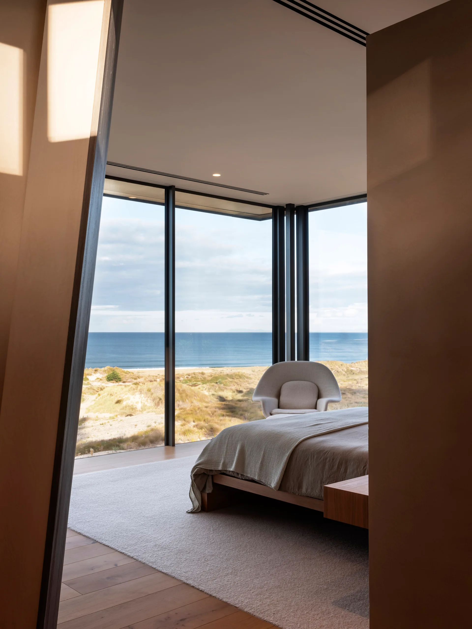 A modern bedroom features a low-profile wooden bed and a curved armchair next to large windows with a coastal ocean view.